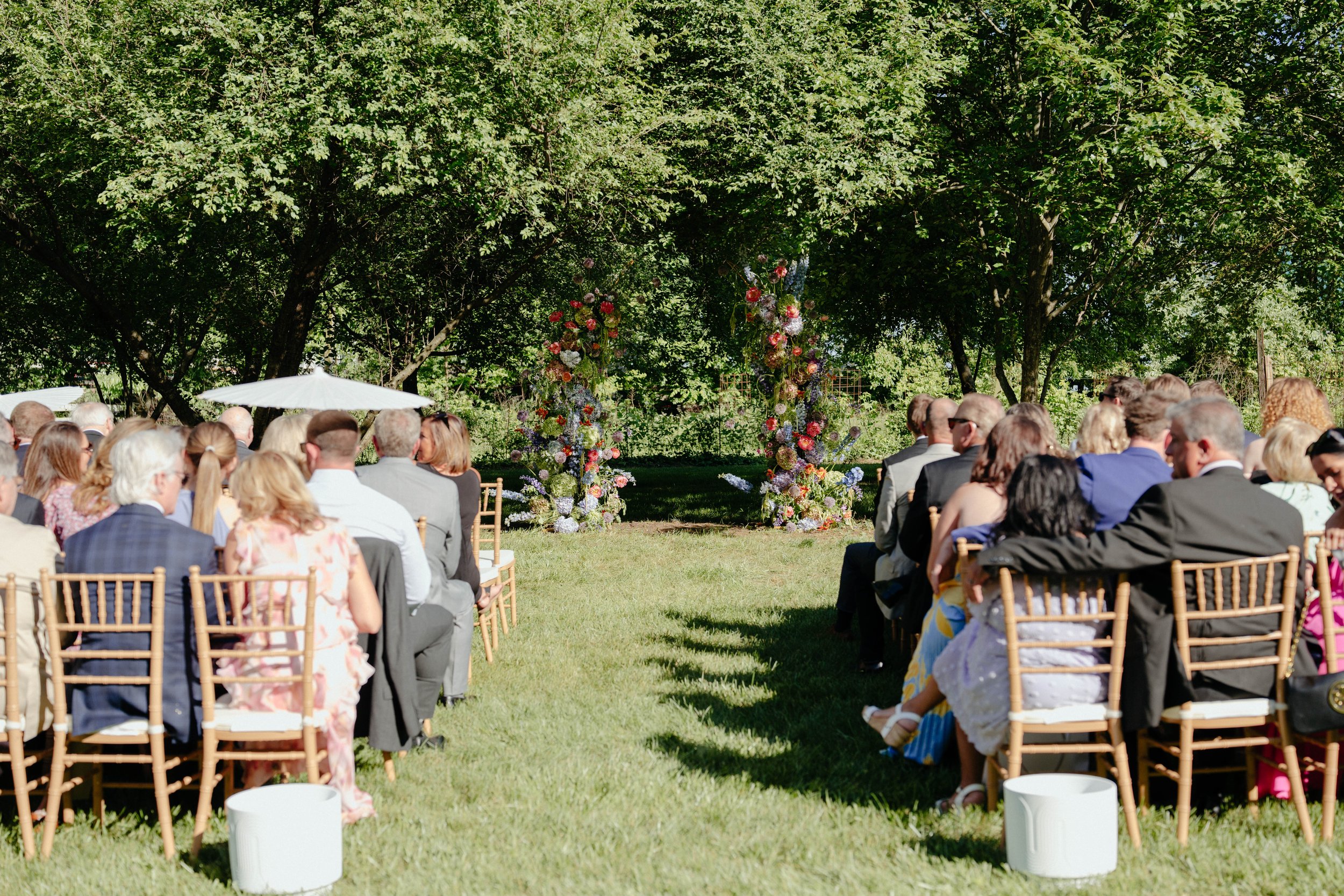Outdoor wedding ceremony with guests seated on wooden chairs facing an arch decorated with flowers, set in a lush green garden with trees in the background.