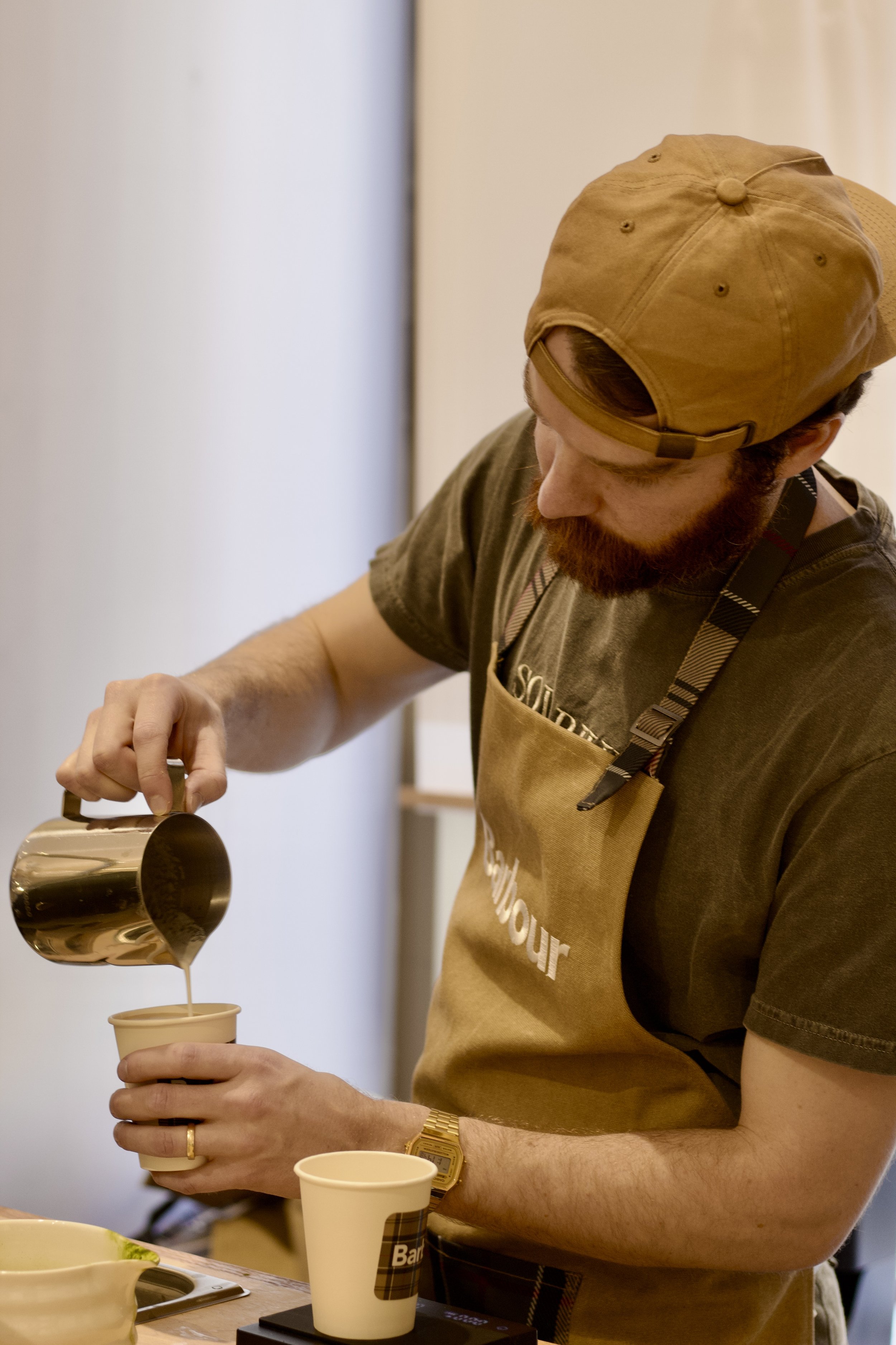 A man wearing a brown apron and a tan cap pours coffee from a metallic jug into a beige paper cup.