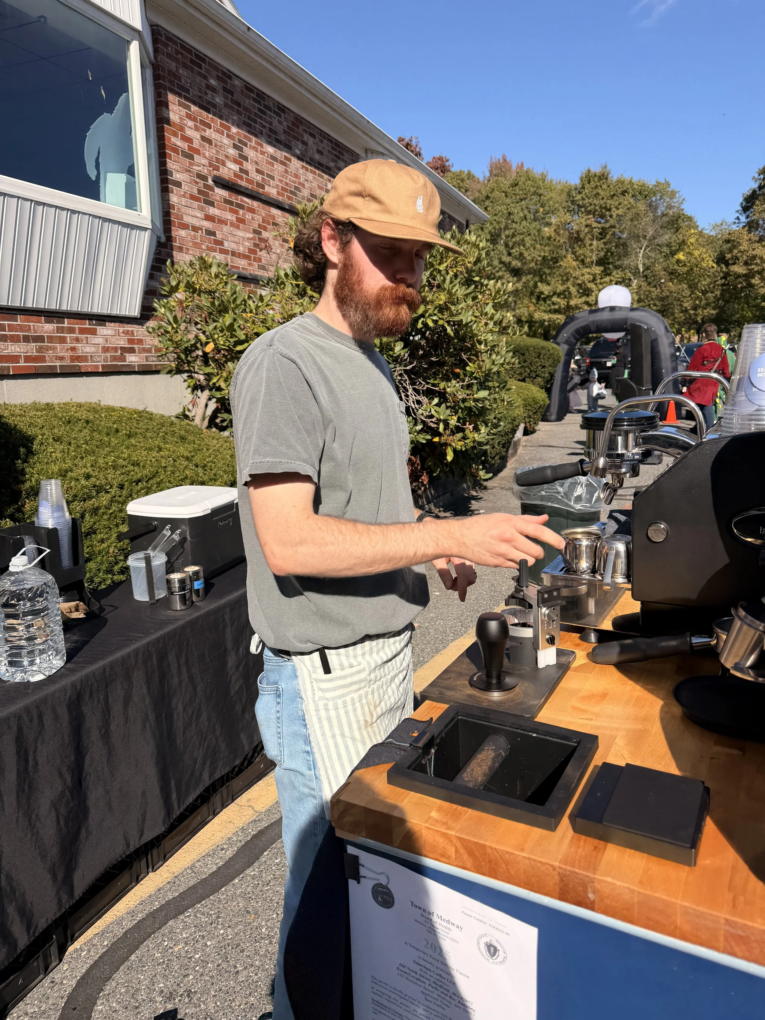 A man with a beard and mustache in a tan cap, gray T-shirt, and striped pants is making coffee at a coffee stand outdoors on a sunny day, with a brick building and trees in the background.