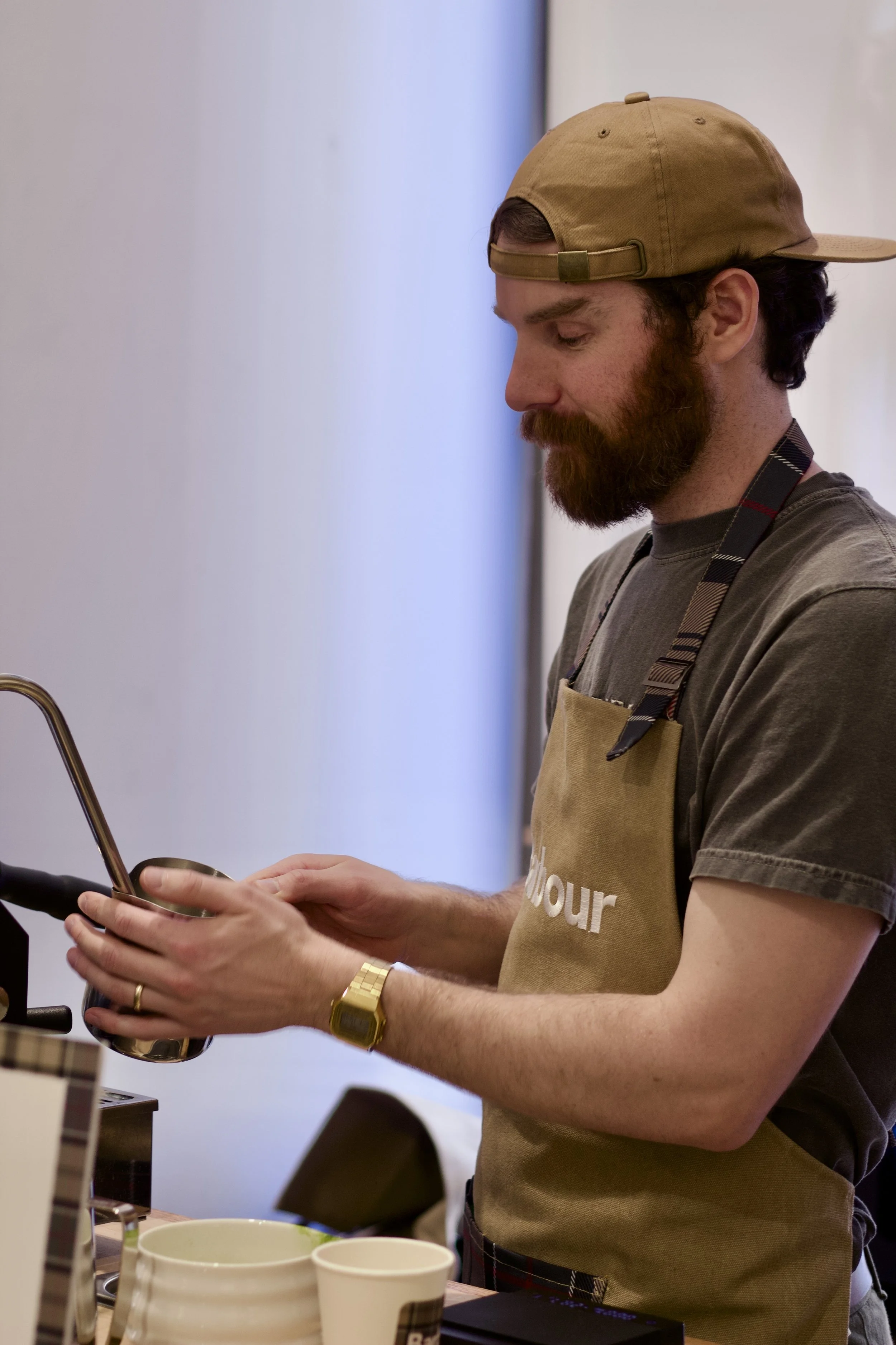 A man with a beard wearing a tan cap, gray t-shirt, gold watch, and an apron, pouring a beverage into a cup at a counter.