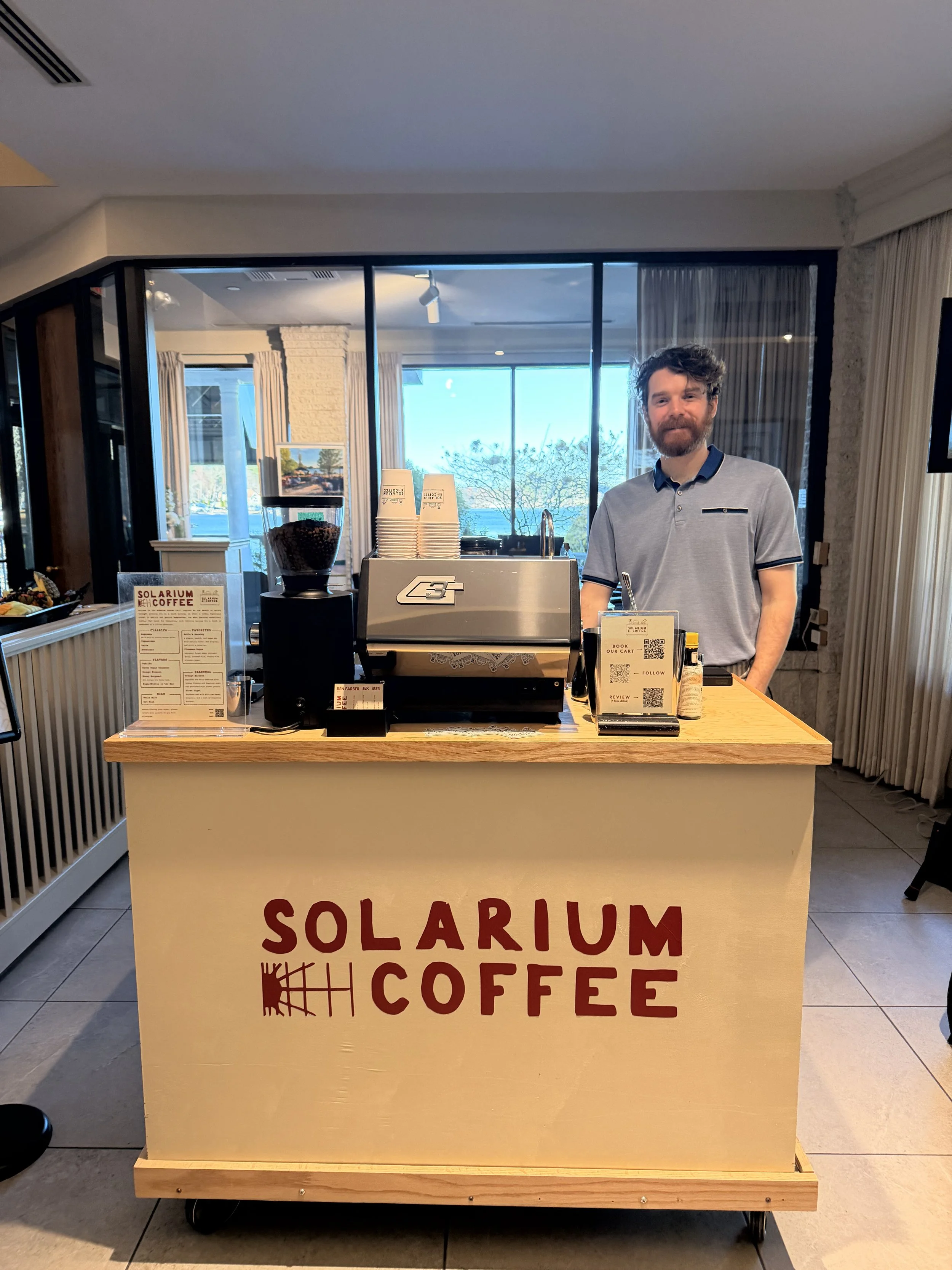 A man standing behind a coffee cart with 'SOLARIUM COFFEE' written on it, in a bright indoor space with large windows revealing a view of trees and a body of water outside.