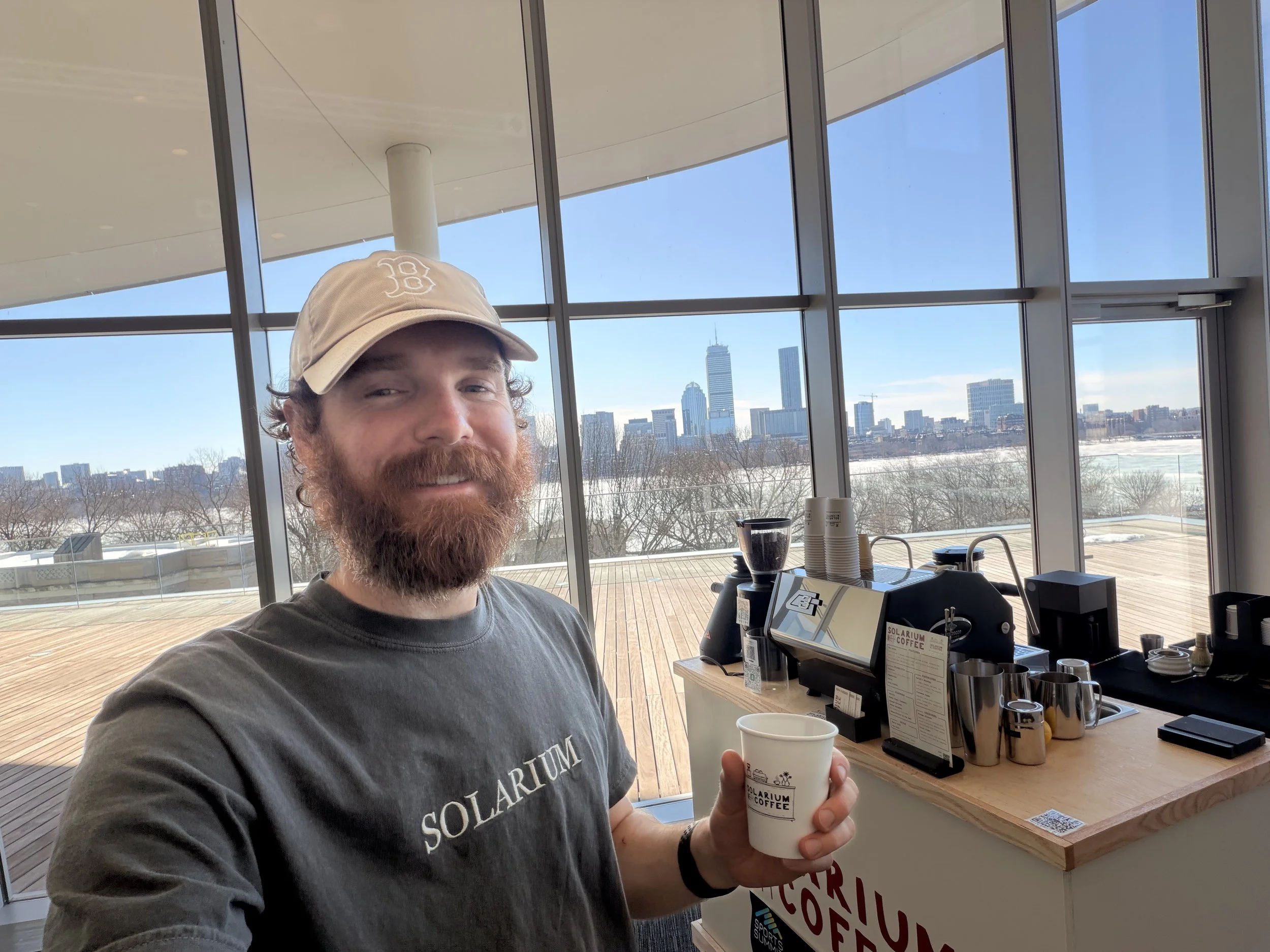 A man with a beard and wearing a beige cap and a gray Solarium t-shirt is holding a white coffee cup. He is standing inside a modern building with large floor-to-ceiling windows showing a city skyline in the background. There is a coffee station on a wooden counter with various coffee-making equipment.