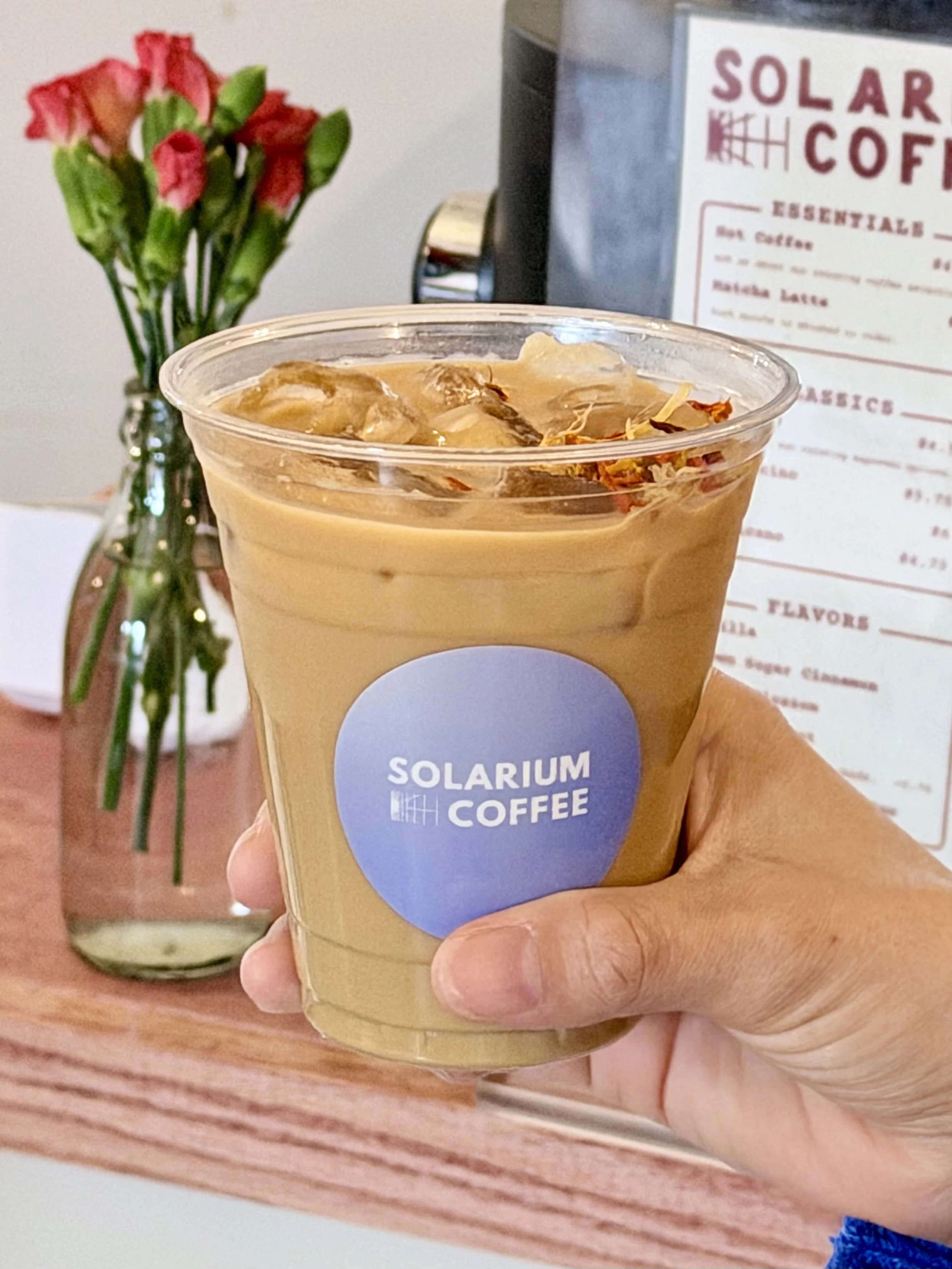 A hand holding a plastic cup of iced coffee with the Solarium Coffee logo, in front of a pink vase with pink flowers and a menu board.
