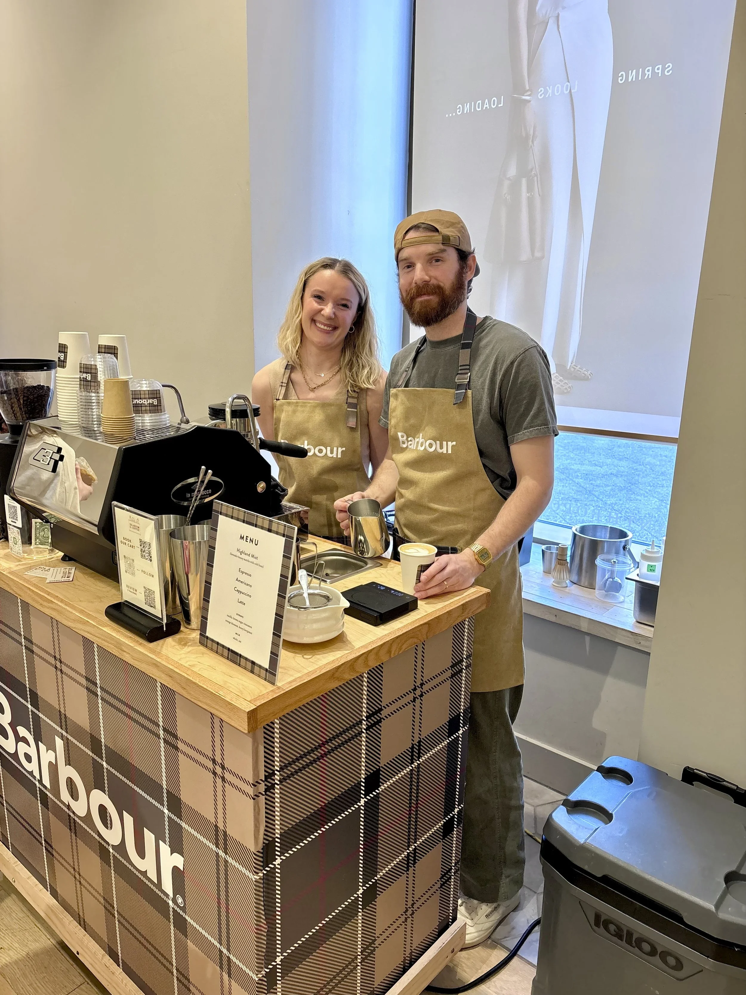 Two baristas wearing aprons smiling behind a coffee counter at Anthropologie on Newbury Street for a Barbour activation with menu, cups, and equipment.