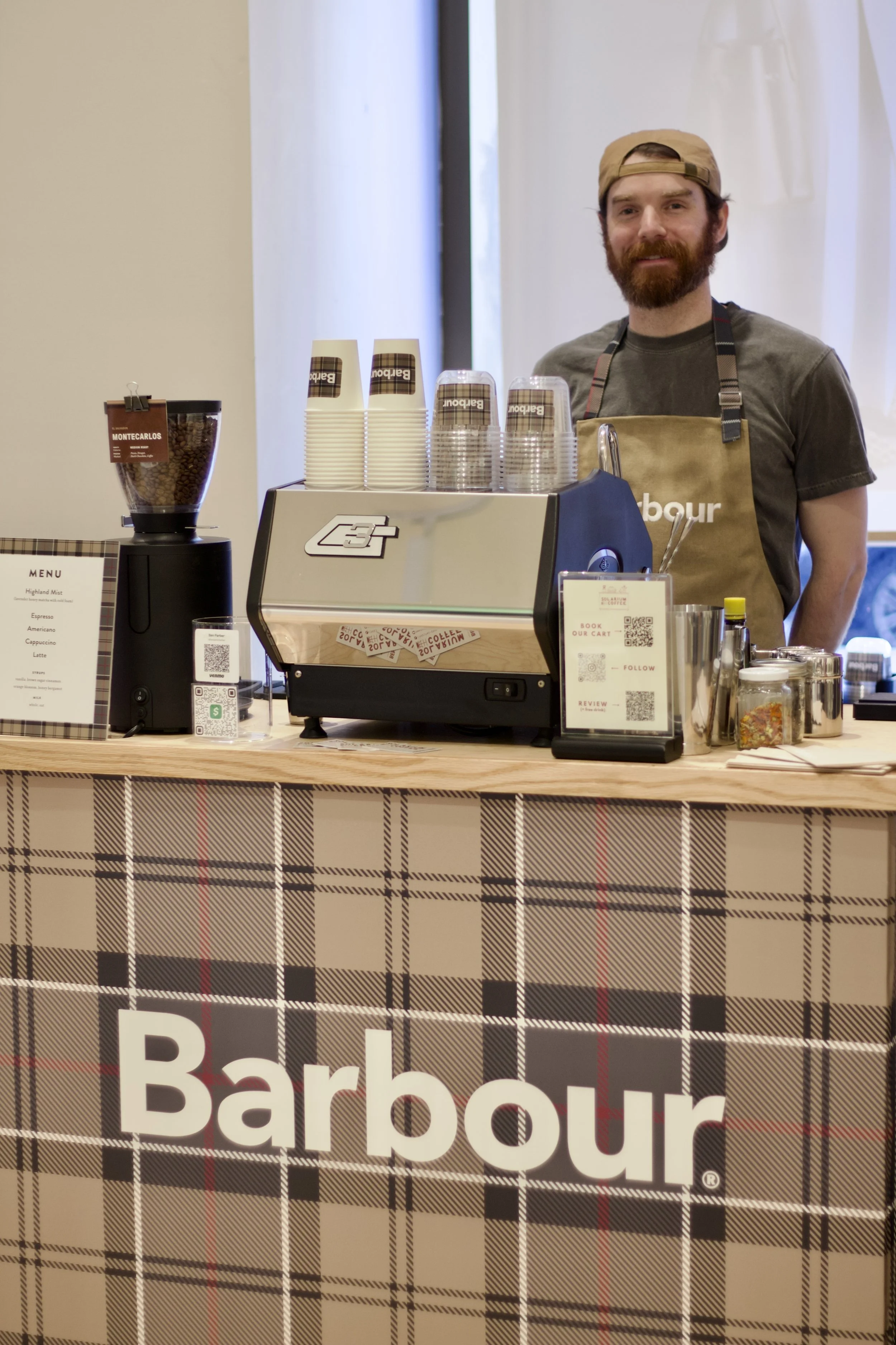 A barista standing behind a coffee counter with a Burberry-patterned front, smiling at the camera, with coffee cups stacked on top of a coffee machine and various jars and equipment on the counter.