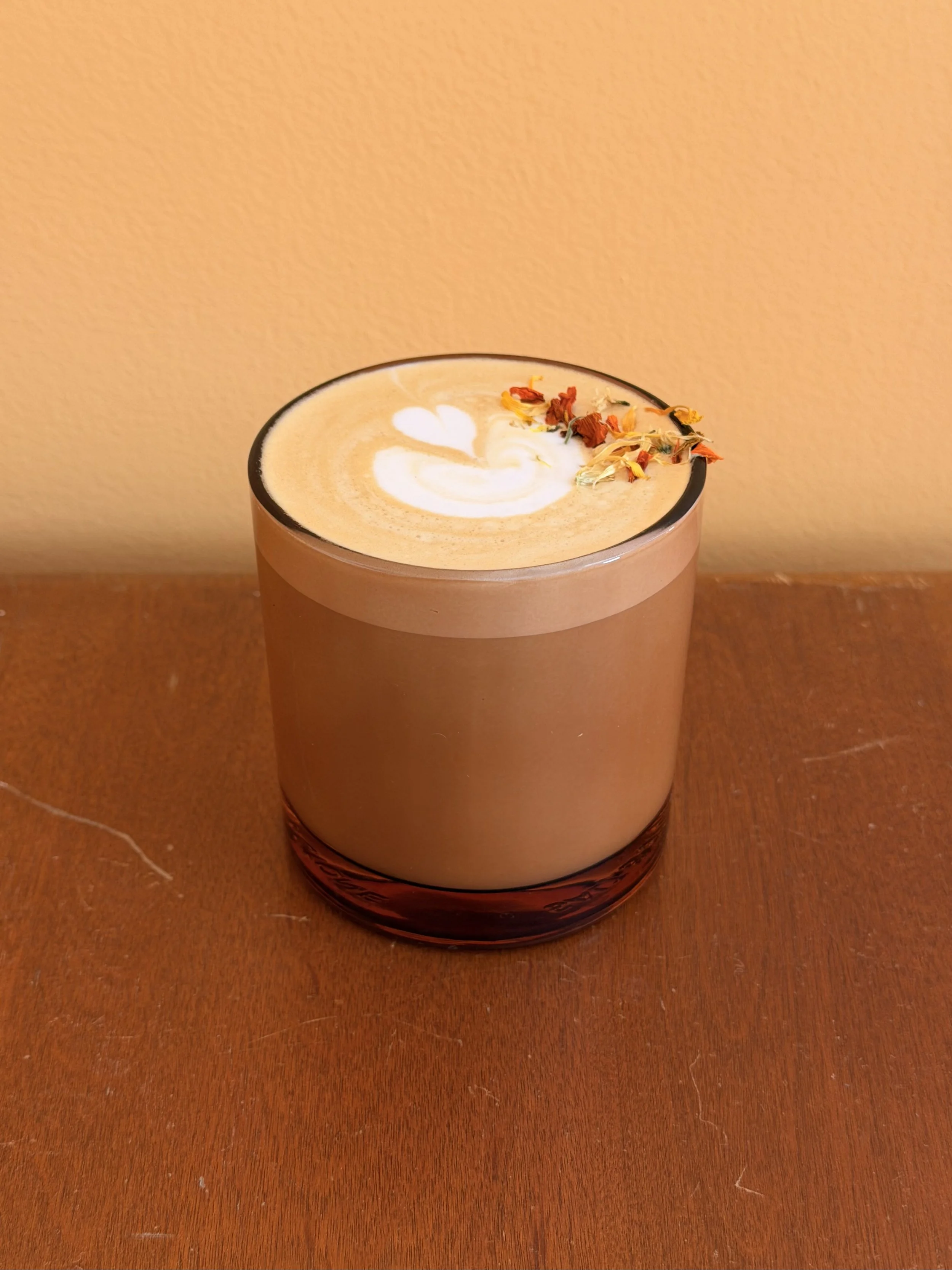 A glass of frothy coffee or latte with latte art and dried flower petals on top, placed on a wooden surface against a plain wall.