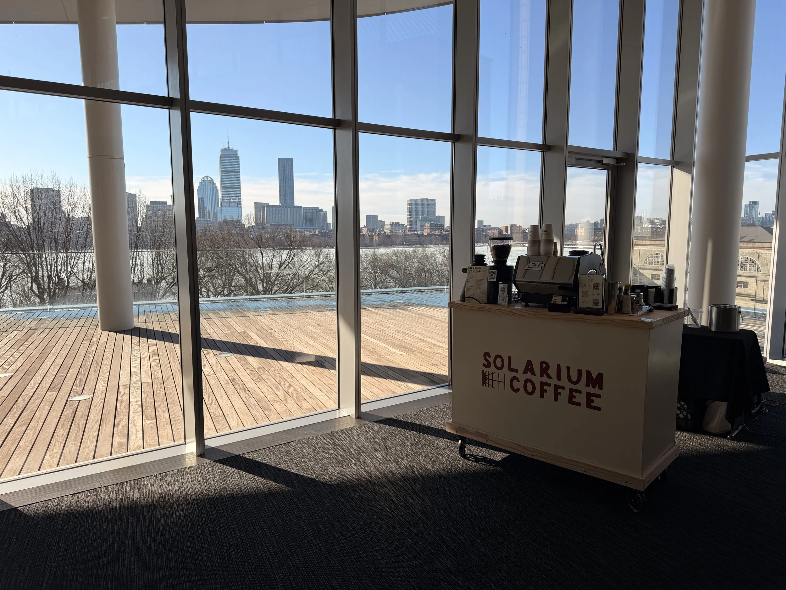Interior of a coffee stand named Solarium Coffee with a city skyline view through large floor-to-ceiling windows, wooden deck outside, and coffee equipment on the counter.