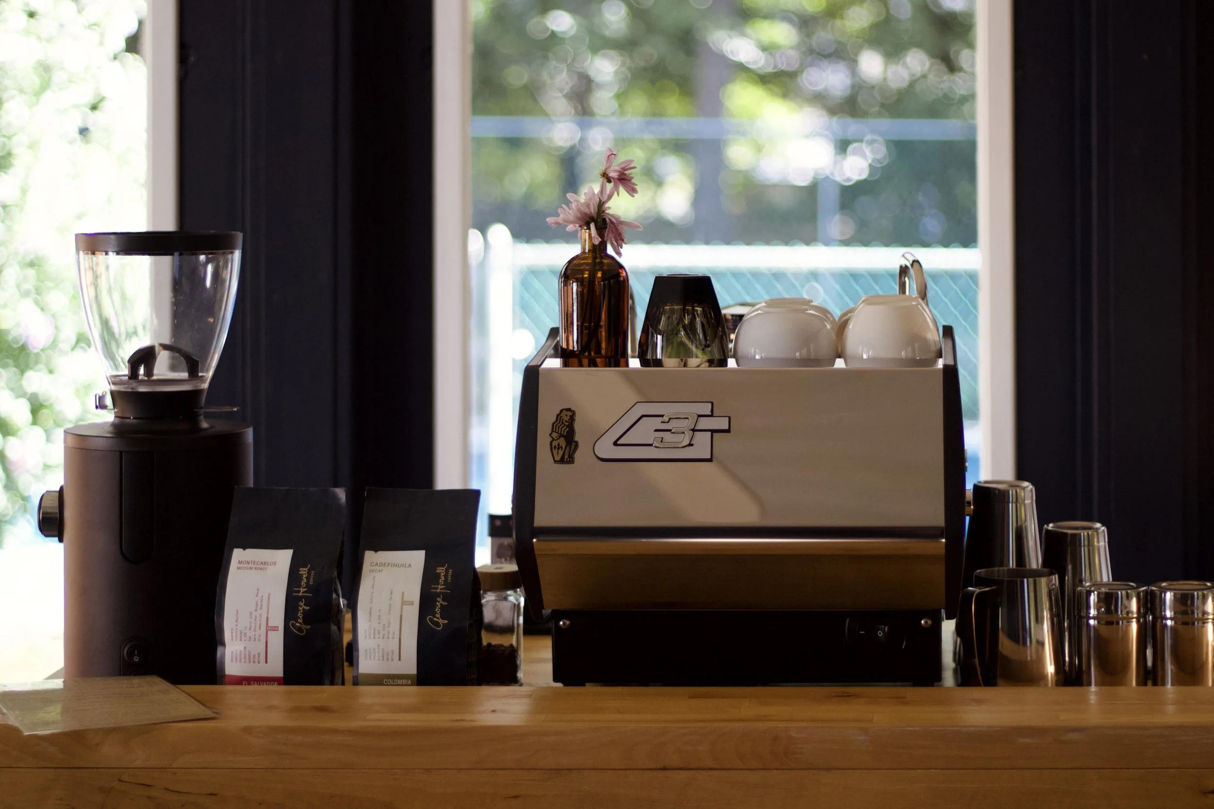 Coffee cart with a coffee grinder, an espresso machine, cups, and coffee bags on a wooden counter, with a window showing an outdoor area in the background.