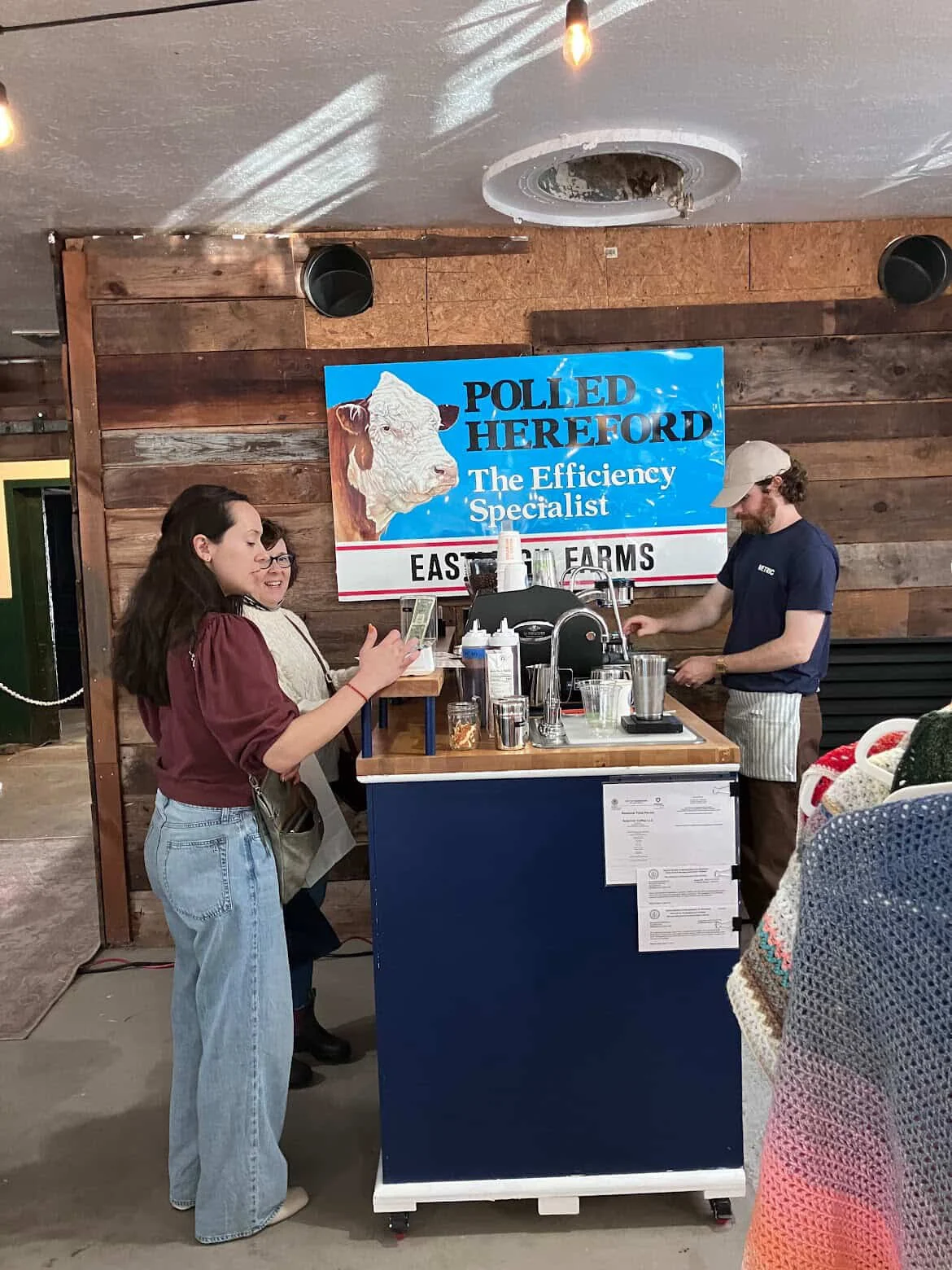 A coffee bar with two women are standing at the counter, one of them holding a money bill, while a man prepares drinks behind the counter. There are various cups, jars, and equipment on the bar.