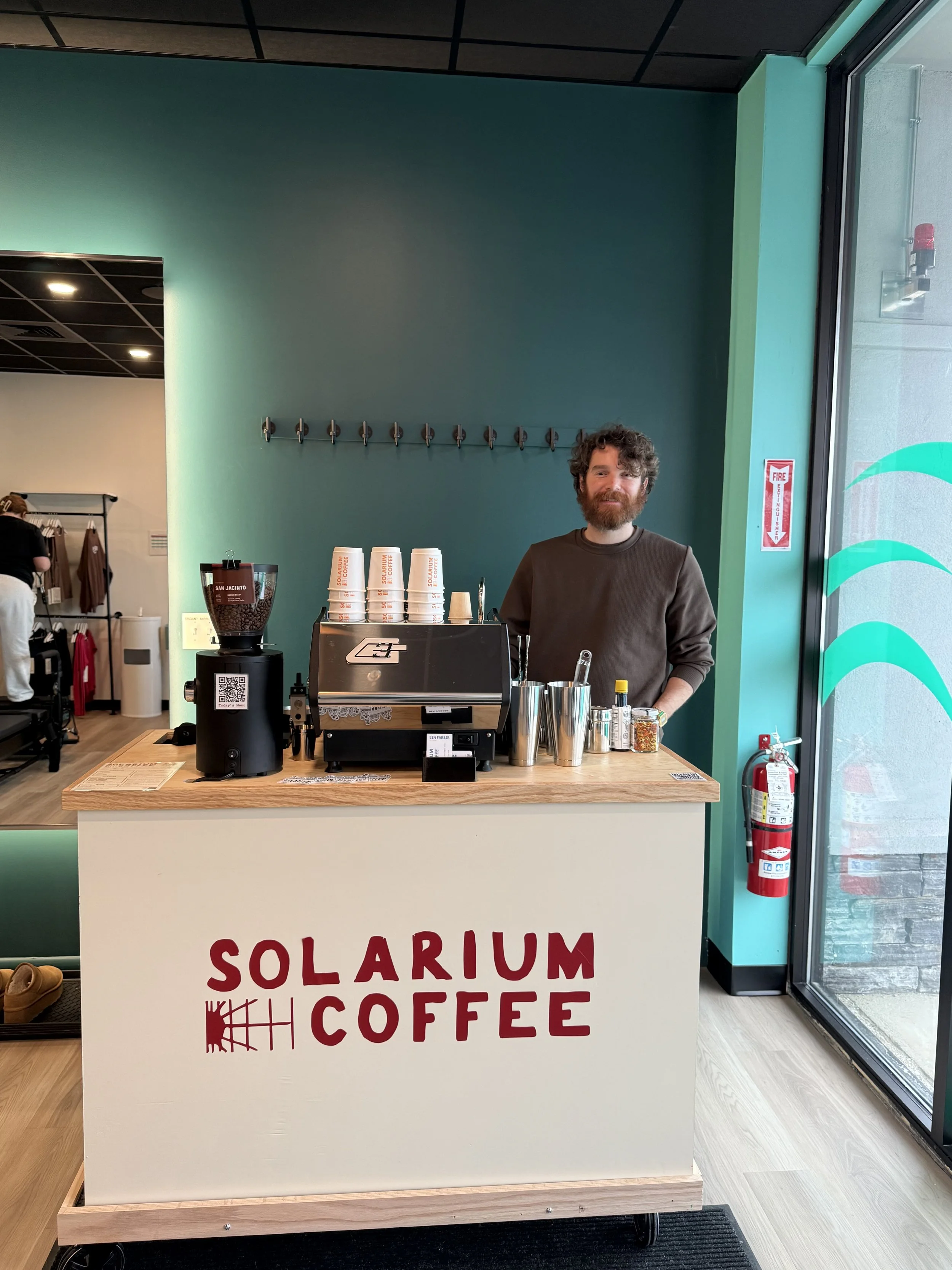 Man standing behind a counter at Solarium Coffee shop, with coffee cups, a coffee machine, and supplies on the counter. The shop has teal and black walls, with a fire extinguisher near the glass door.