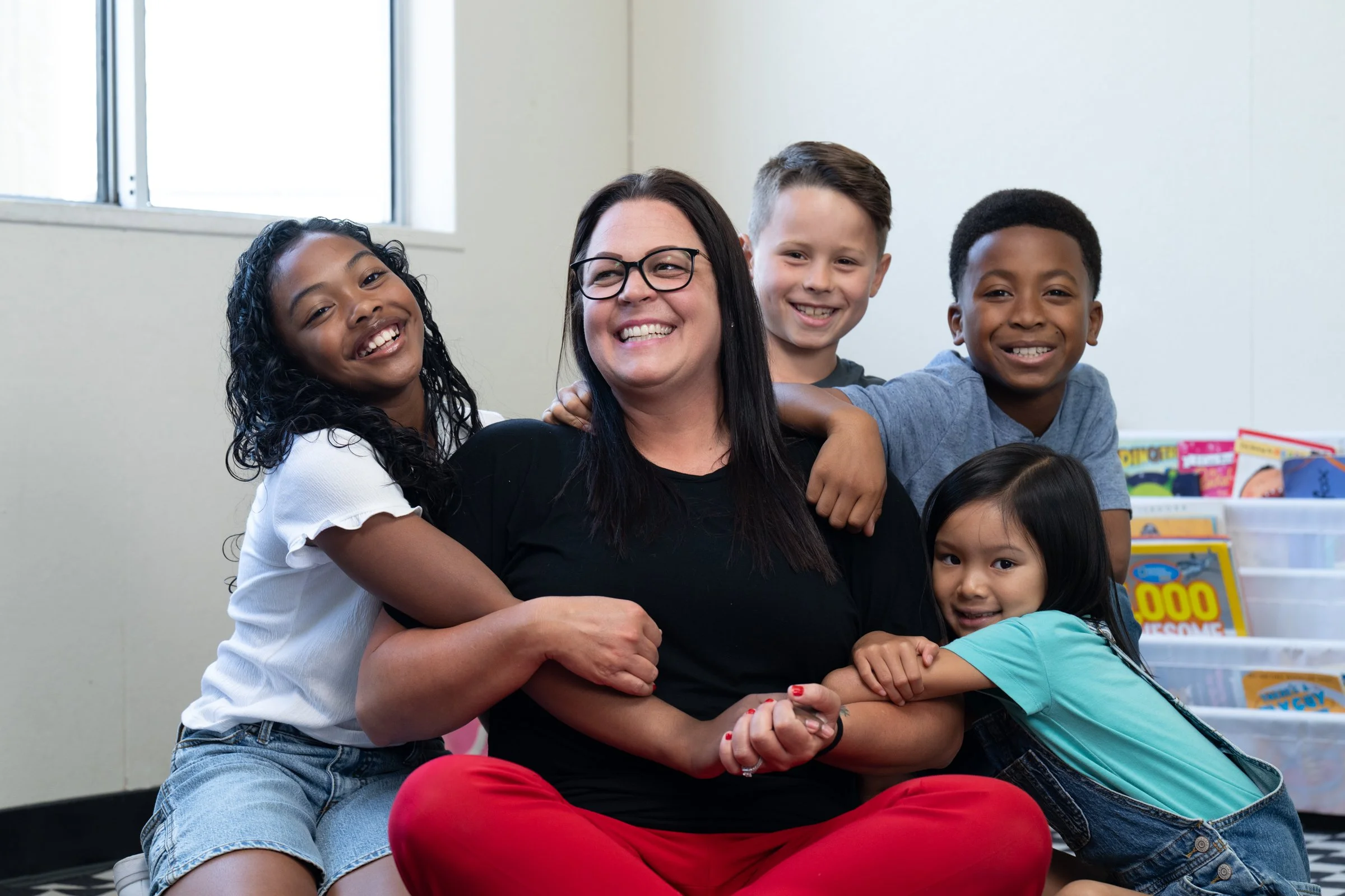 A smiling woman surrounded by four children, all sitting together in a classroom with bookshelves in the background.