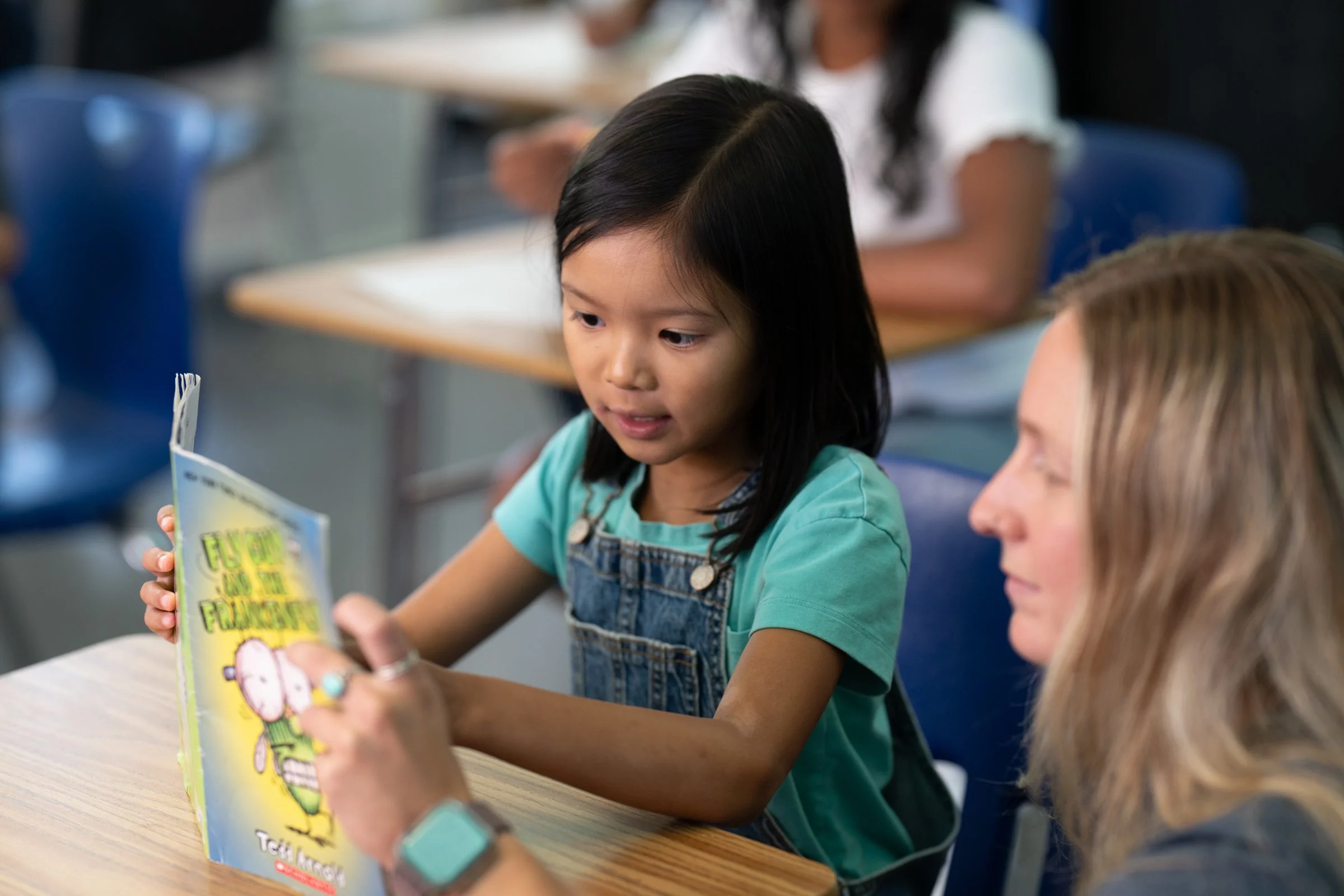A young girl with black hair wearing a teal shirt and denim overall, reading a colorful book titled 'The Sheep in the Fluffiest,' while an adult woman with blonde hair looks on. They are seated at a wooden table in a classroom setting.
