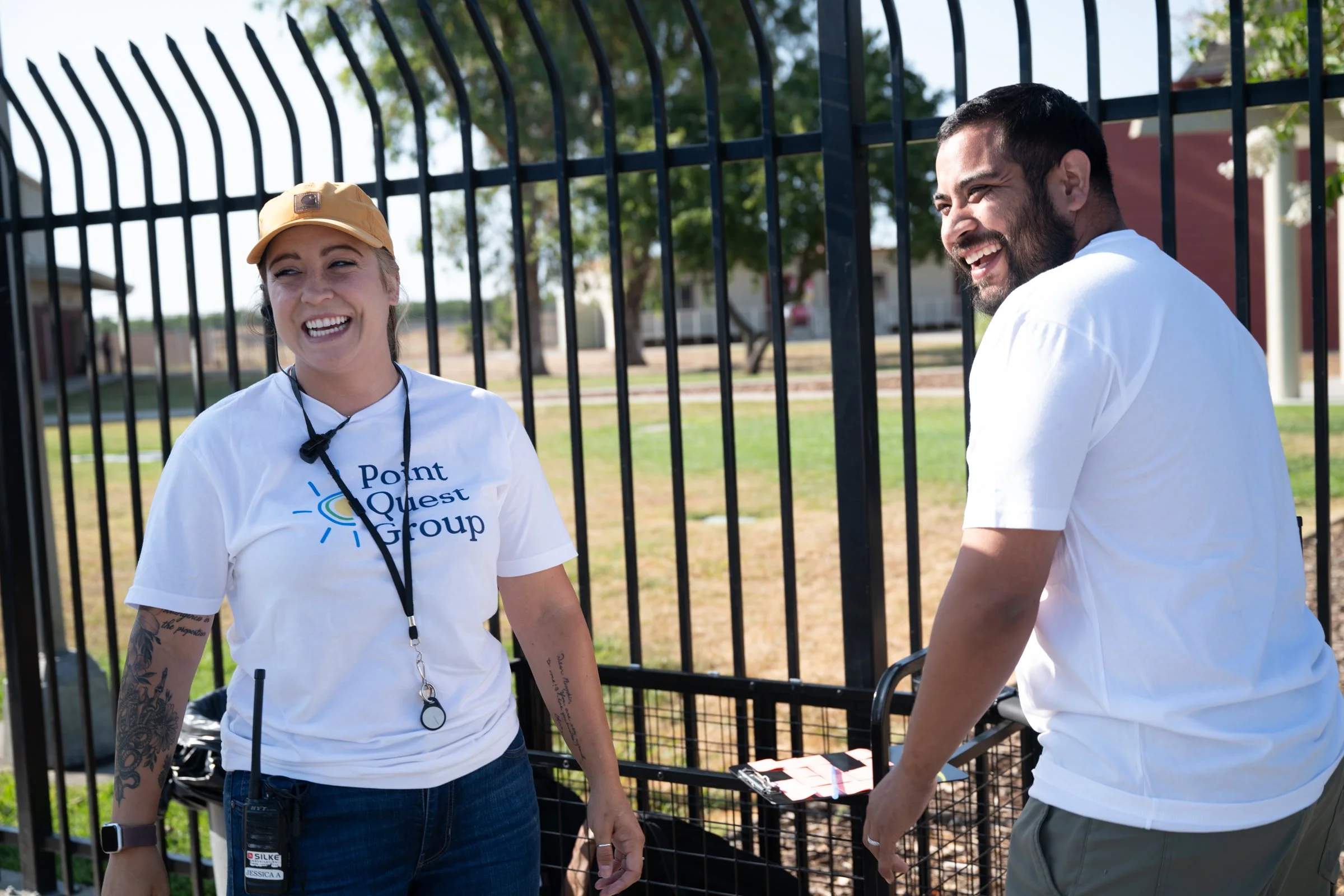 Two people are standing and smiling outdoors near a black metal fence, with trees and houses in the background. They are wearing white t-shirts, and one has a yellow cap and a walkie-talkie. They appear to be having a friendly conversation.