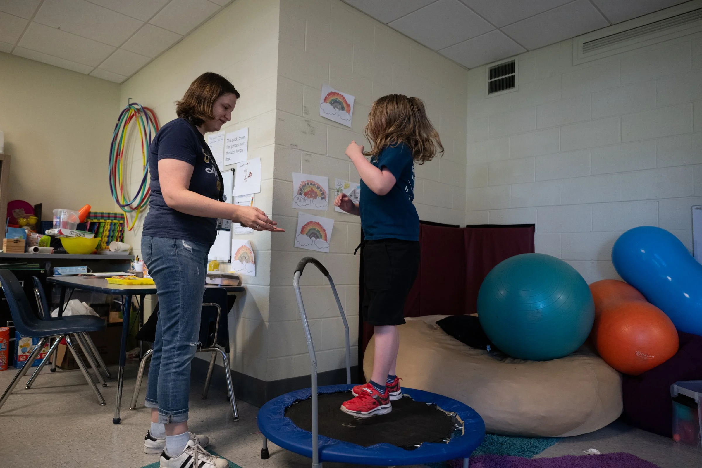 A woman assisting a young girl practicing on a mini trampoline in a therapy or classroom setting, with colorful therapy balls and wall decorations featuring rainbows.