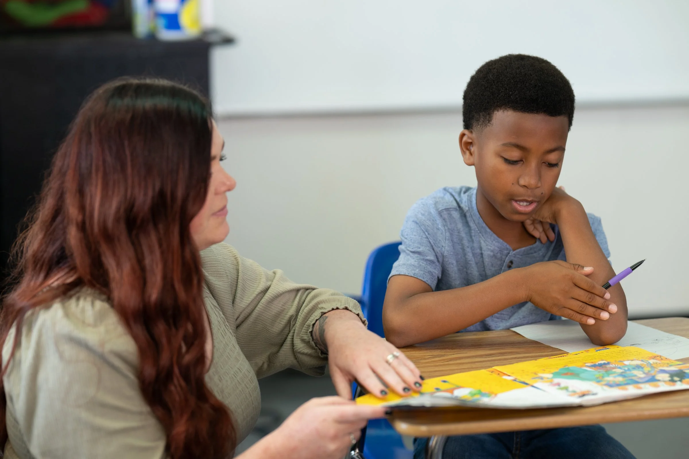 A woman and a teenage boy studying together at a wooden table in a library, looking at a laptop.