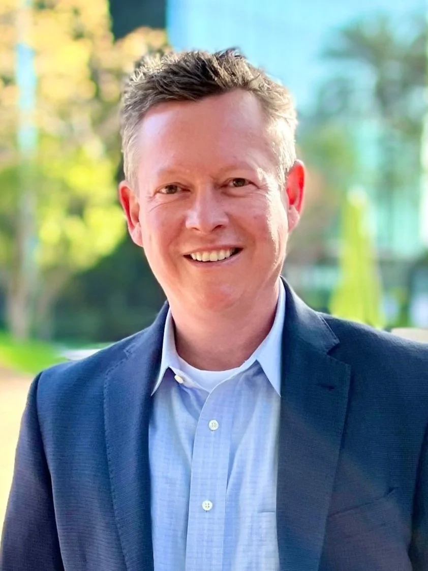 Smiling man in a navy blazer and light blue collared shirt outdoors with blurred green trees in the background.