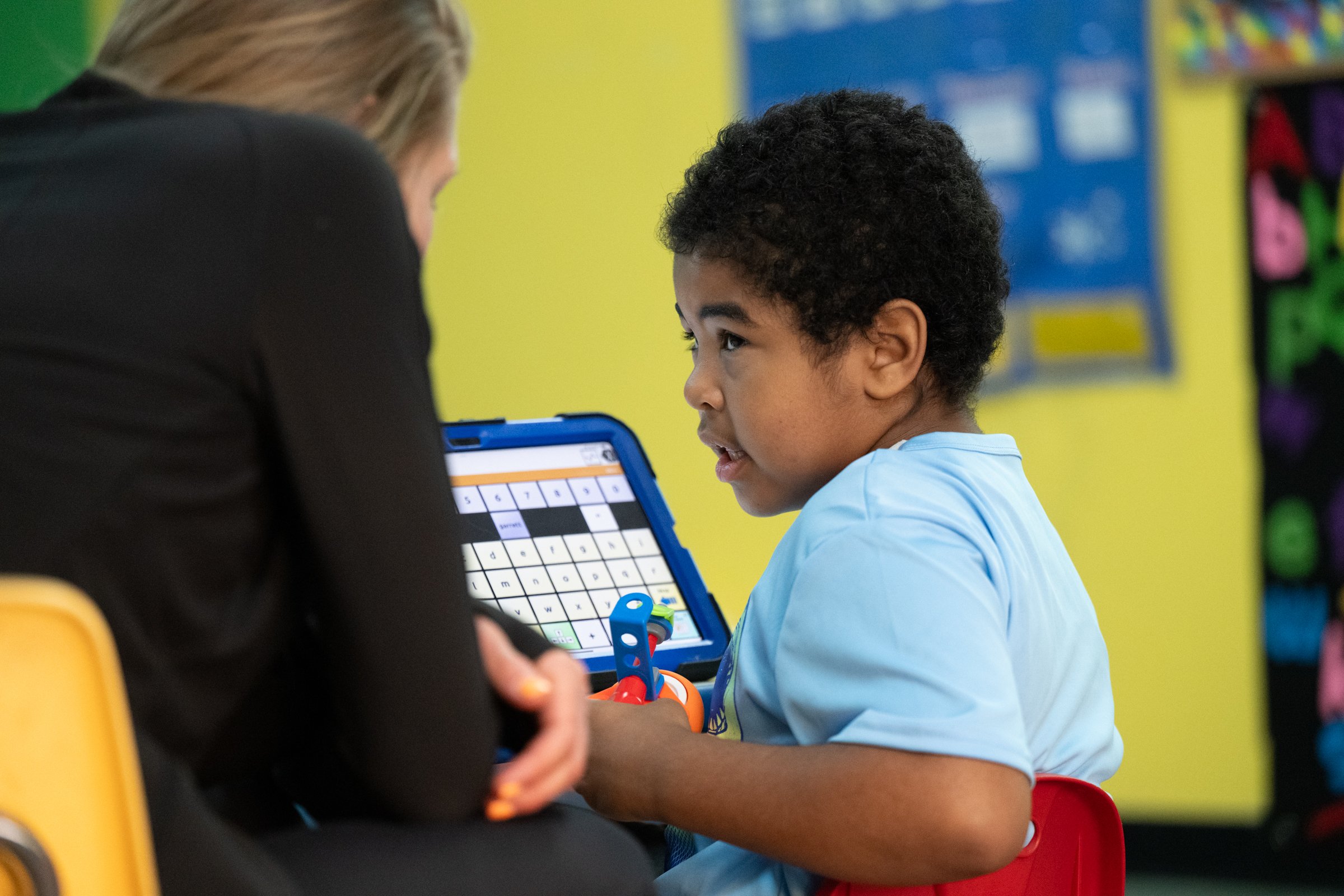 A young boy and an adult woman are engaged in an activity using a tablet, which displays a crossword puzzle. The boy has curly black hair and is wearing a light blue shirt. The background shows colorful classroom decorations.