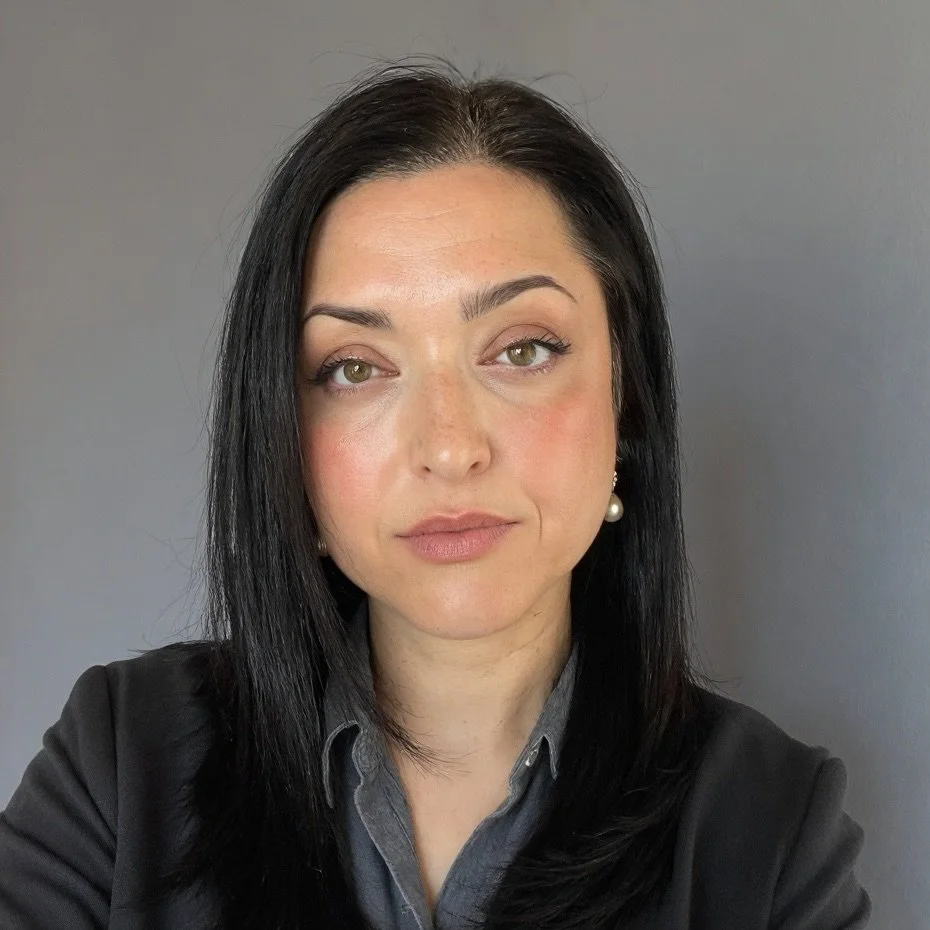A woman with dark hair and light eyes, wearing a grey collared shirt, black jacket, and a pearl earring, standing against a plain grey background.