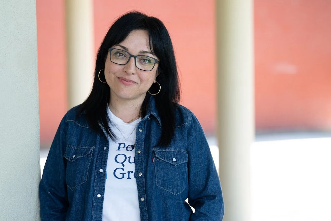 Woman with black hair and glasses standing outdoors near a painted wall, wearing a denim jacket and white t-shirt.