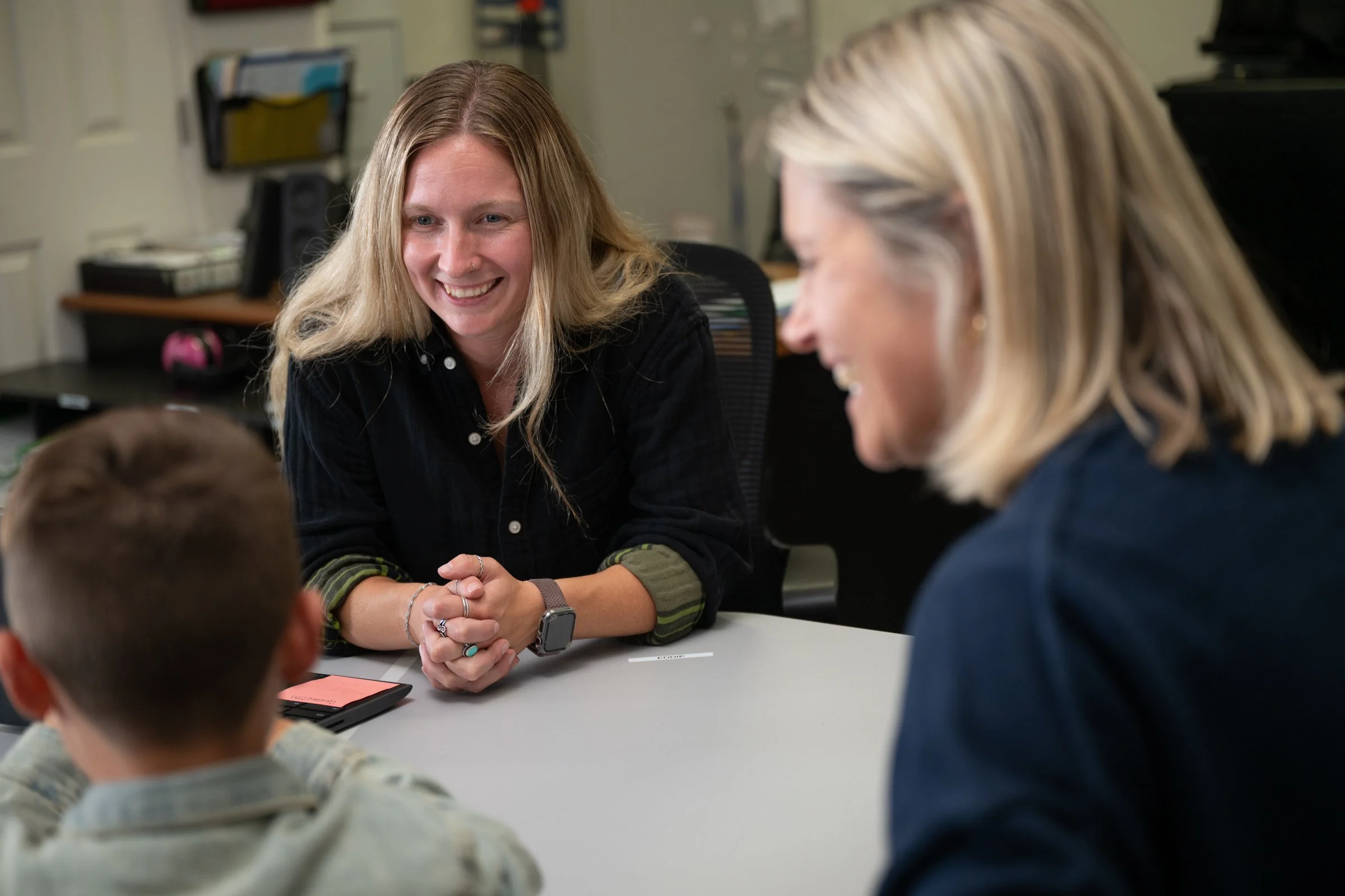 Two women and a young boy sitting around a table, smiling and engaging in conversation in an office environment.