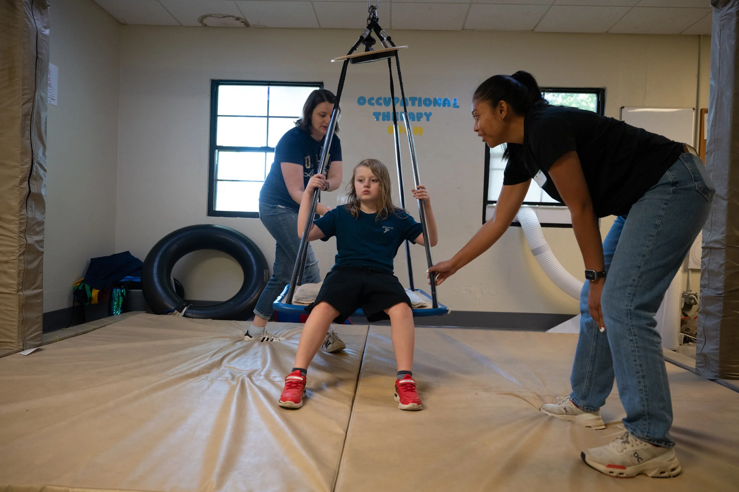 Child sitting on a swing in an occupational therapy room with two adults assisting. The room has windows, therapy equipment, and a sign on the wall that reads 'Occupational Therapy'.