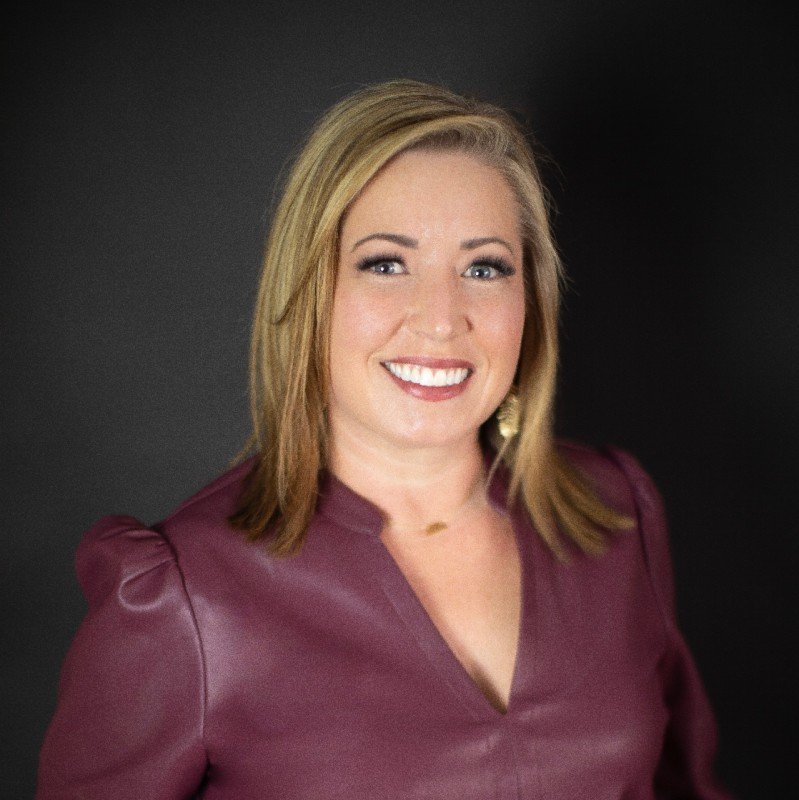A woman with shoulder-length blonde hair, smiling, wearing a dark maroon blazer against a black background.