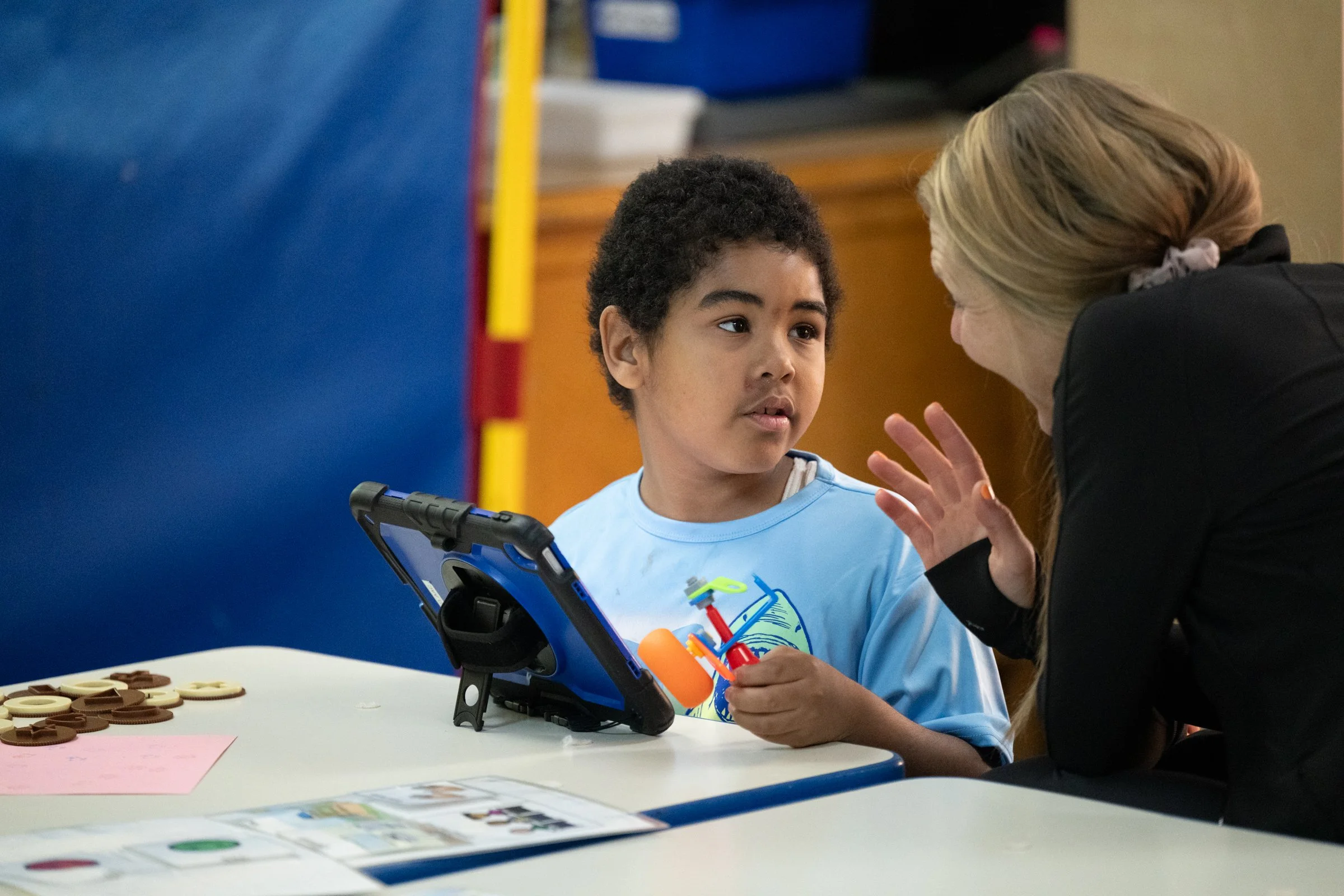 A young boy sits at a table, talking to a woman who appears to be explaining something to him. The boy has curly hair and is holding a small object. There are chocolate tokens and sheets of paper on the table, and a tablet device propped up in front of him.