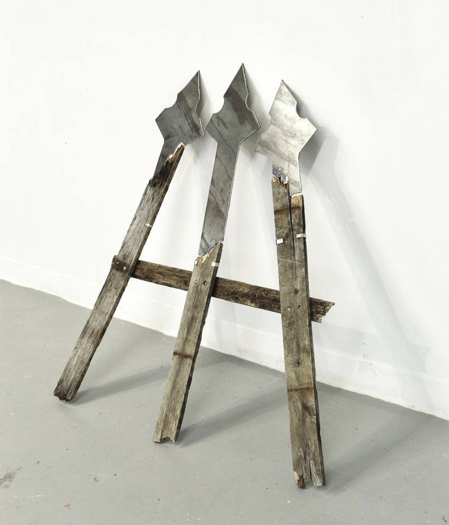 A minimalist sculpture made of wood and steel that resembles three pointed fence posts aimed at a point.