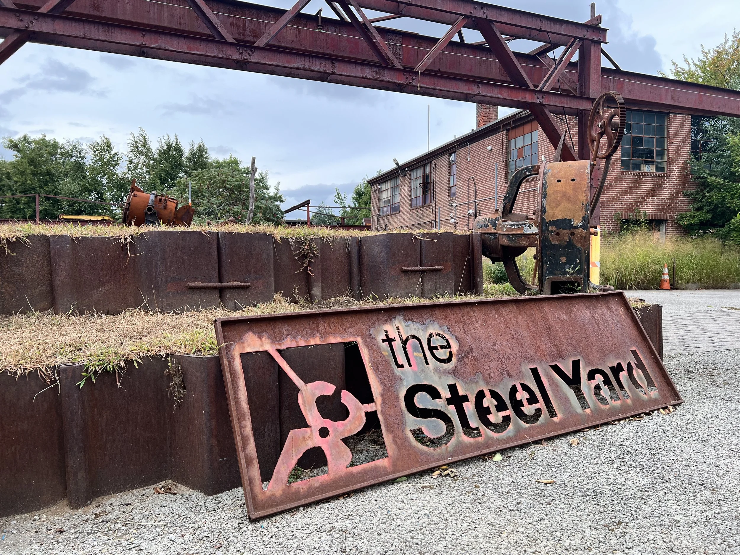 An outdoor view of a metal sign that reads "the steel yard" pictured in a courtyard under steel girders.