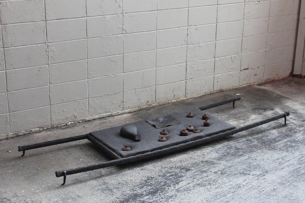 A black mixed media sculpture that resembles a stretcher on the ground in a concrete room