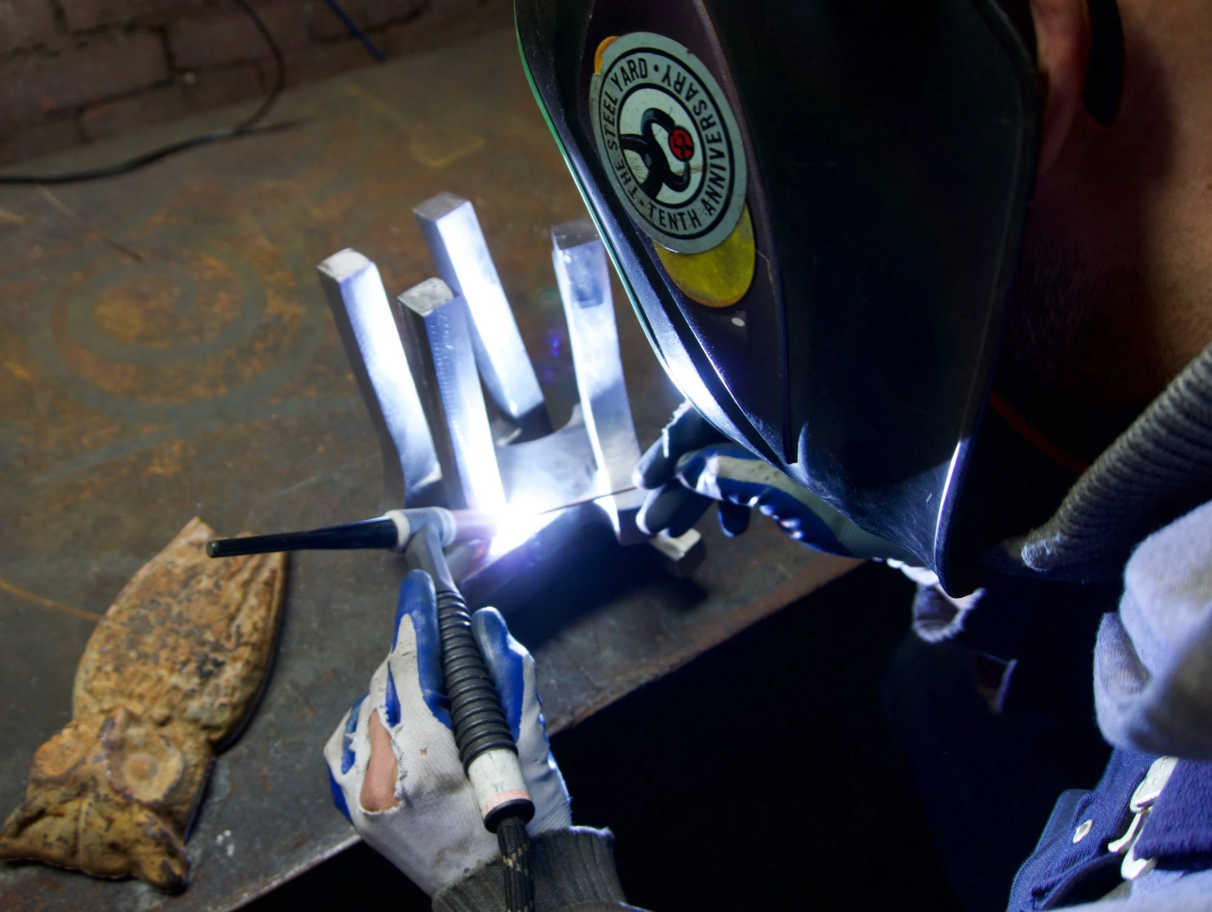 A tight view of a person welding an abstract steel sculpture. The figure is seen from behind and the welding is pictured over their shoulder.