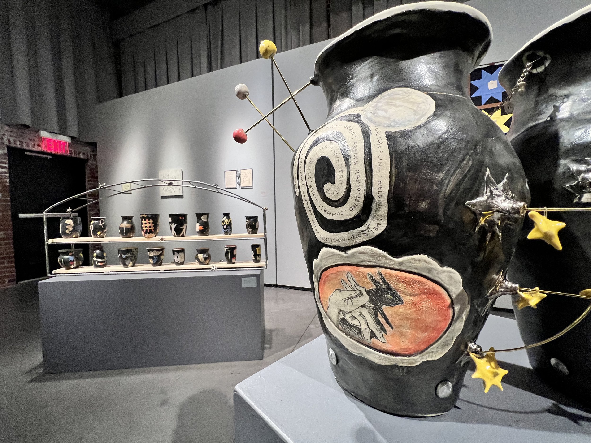 A view into an art gallery featuring a large black vase with illustrations in the foreground and a collection of smaller black vases with illustrations in the background.