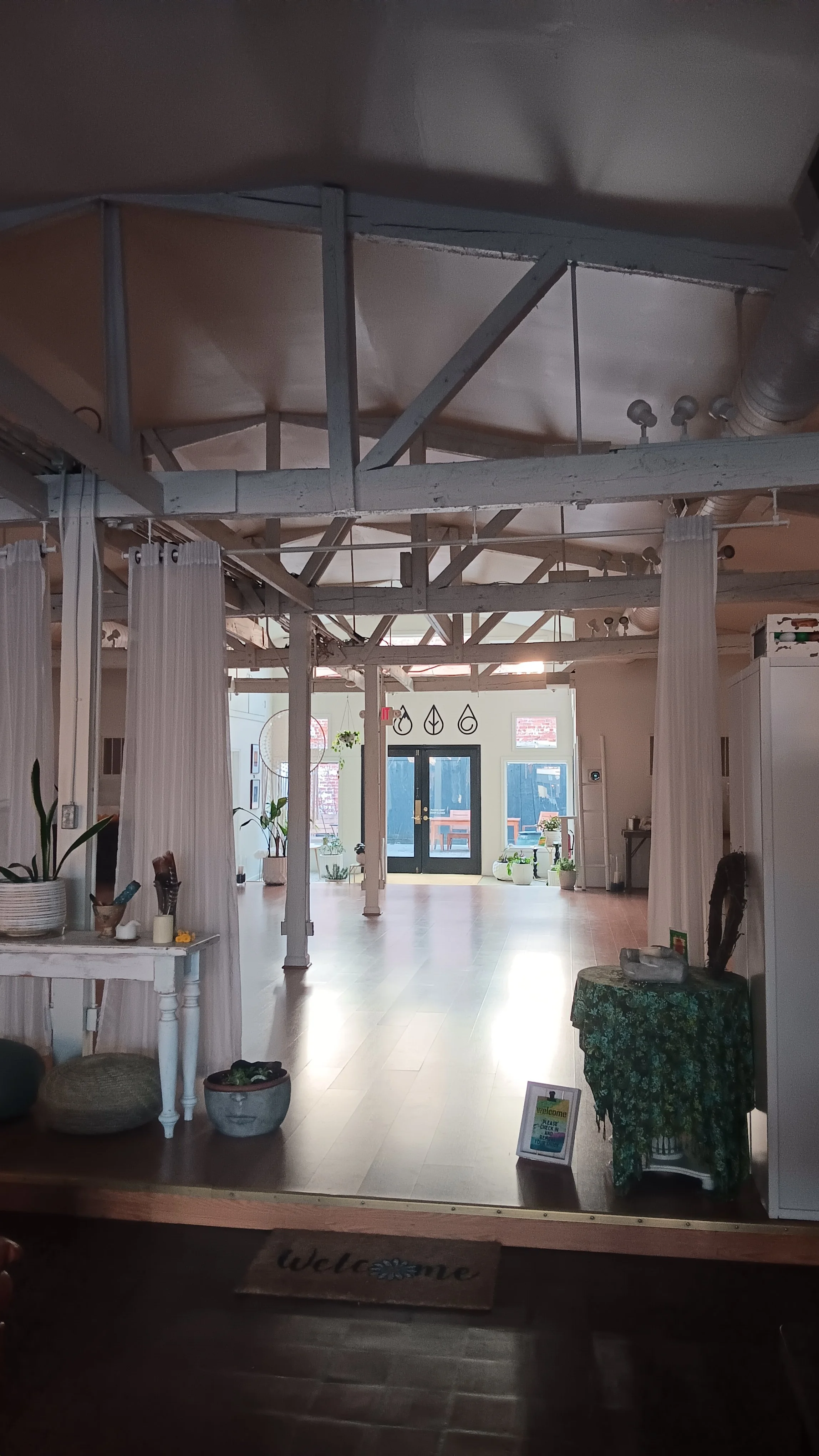 Interior of a spacious, light-filled yoga studio with white walls and wooden floors. Decor includes hanging art symbols, numerous potted plants, white curtains, and a welcoming sign at the entrance that says 'Welcome.'