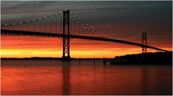A suspension bridge at sunset over a body of water, with orange and pink sky and illuminated cables.