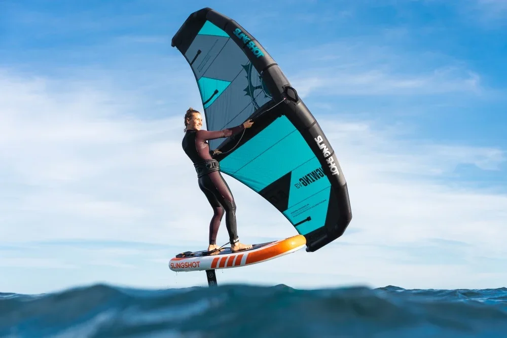 Person flying on a wing suit attached to a hydrofoil board over the water against a blue sky.