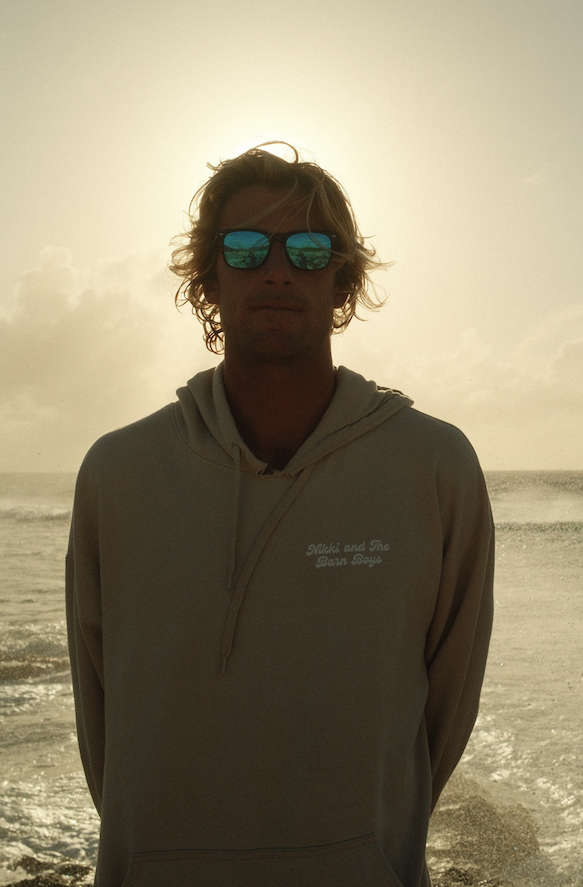 A person with wavy hair wearing sunglasses and a hoodie standing on a beach during sunset.