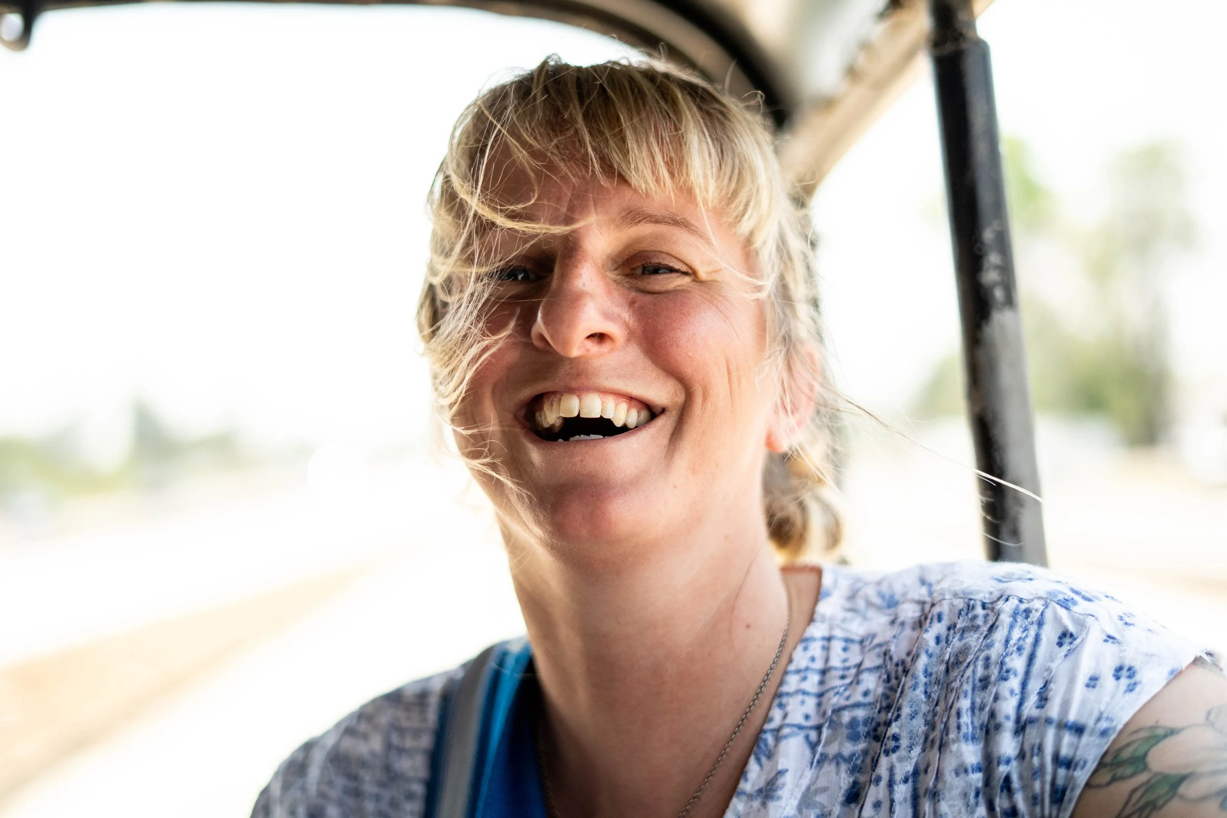 A woman with blonde hair smiling openly, showing her teeth, outdoors in a bright setting.