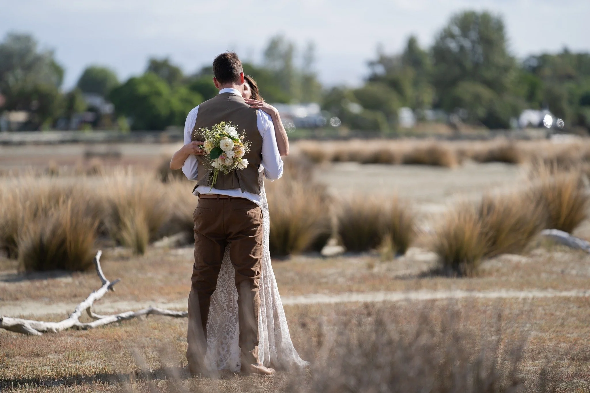 A couple embracing outdoors, with the man holding a bouquet of white flowers on his back, in a natural landscape with grass and bushes.