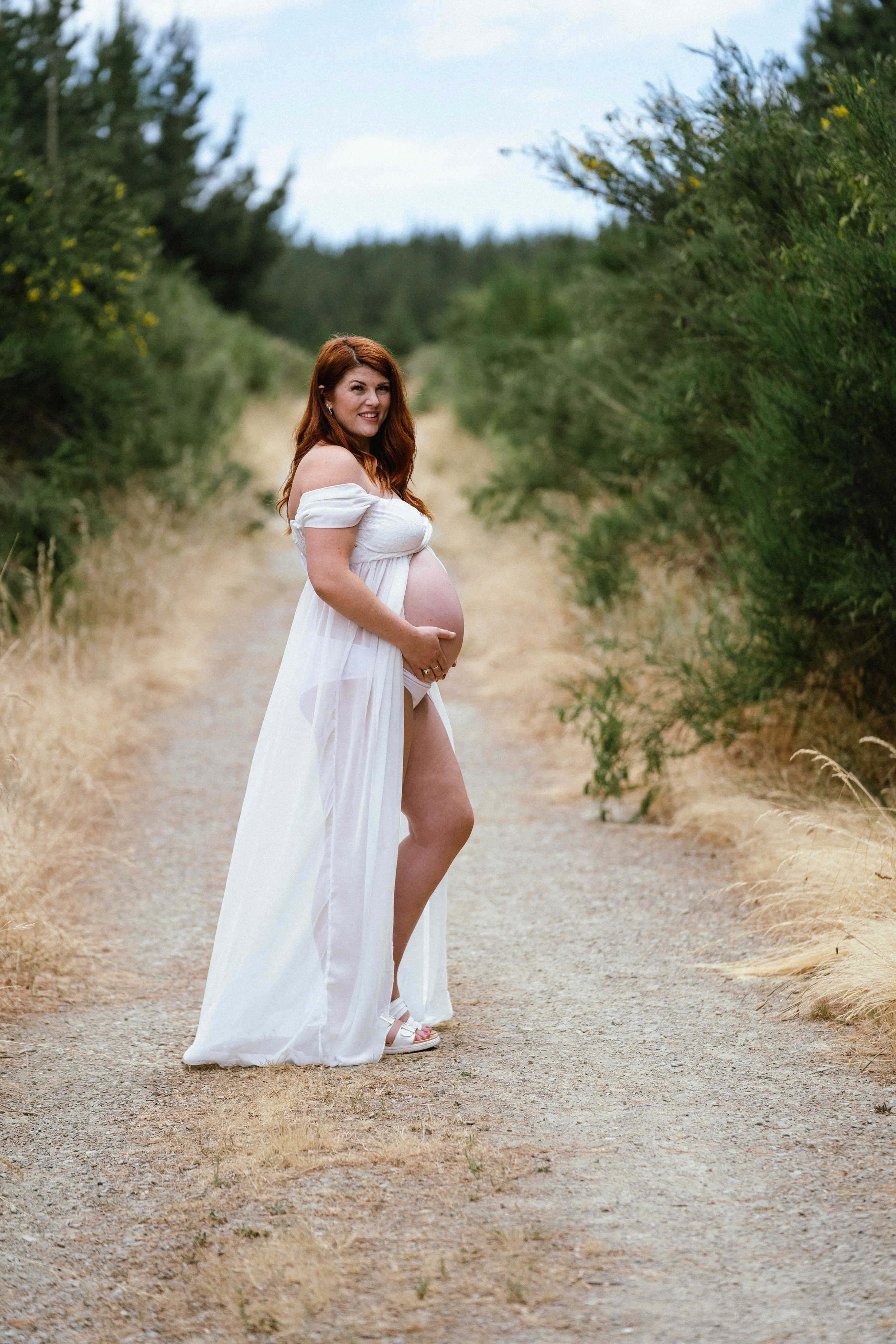 A pregnant woman with red hair in a white flowing dress standing on a dirt path surrounded by greenery, smiling and holding her belly.