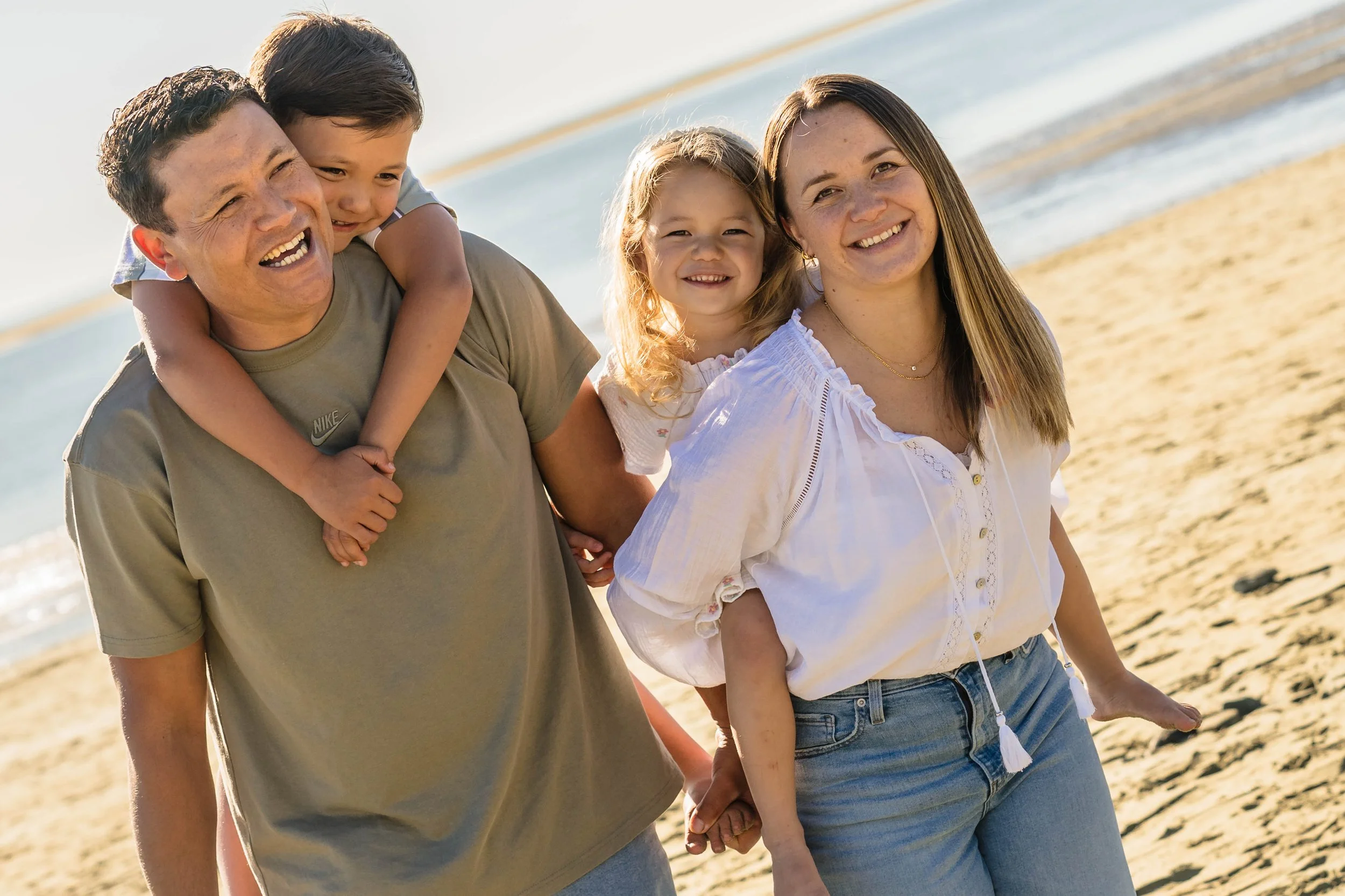 A happy family of four with two adults and two children walking on a sandy beach, holding hands and smiling.
