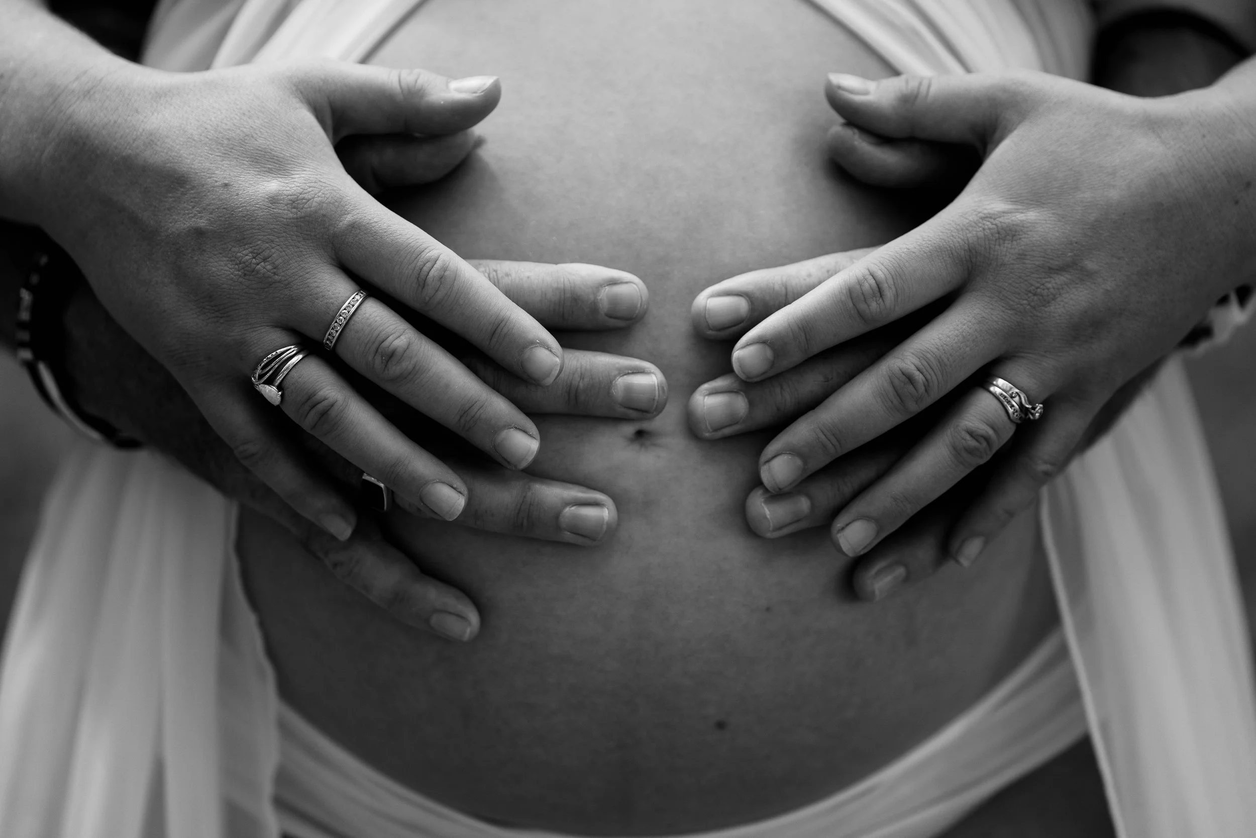 Black and white photograph of a pregnant woman's belly with two pairs of hands placed on it, one on each side, with visible rings on the fingers.