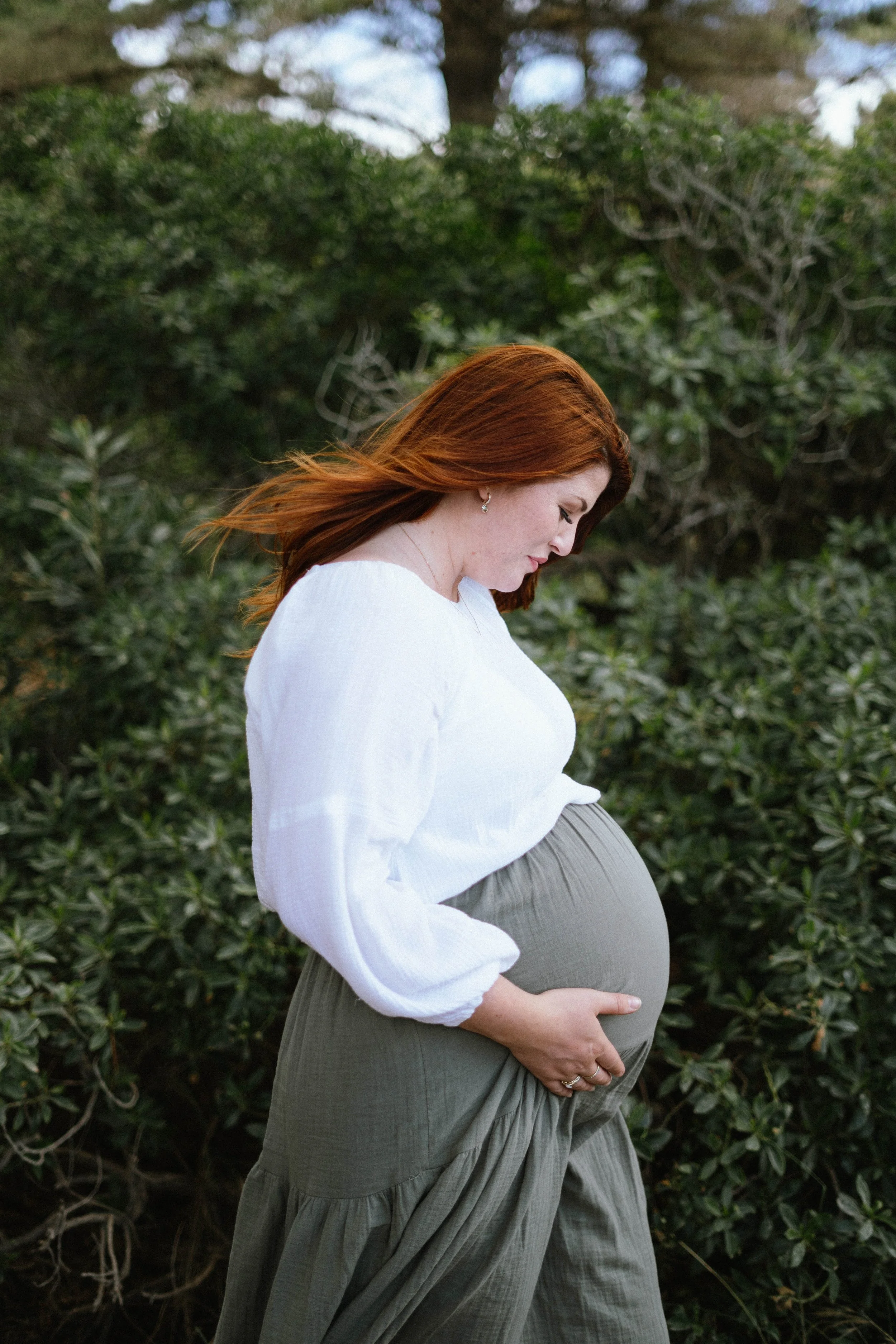 A pregnant woman with red hair, wearing a white long-sleeve top and olive green pants, standing outdoors among greenery, gently holding her baby bump.