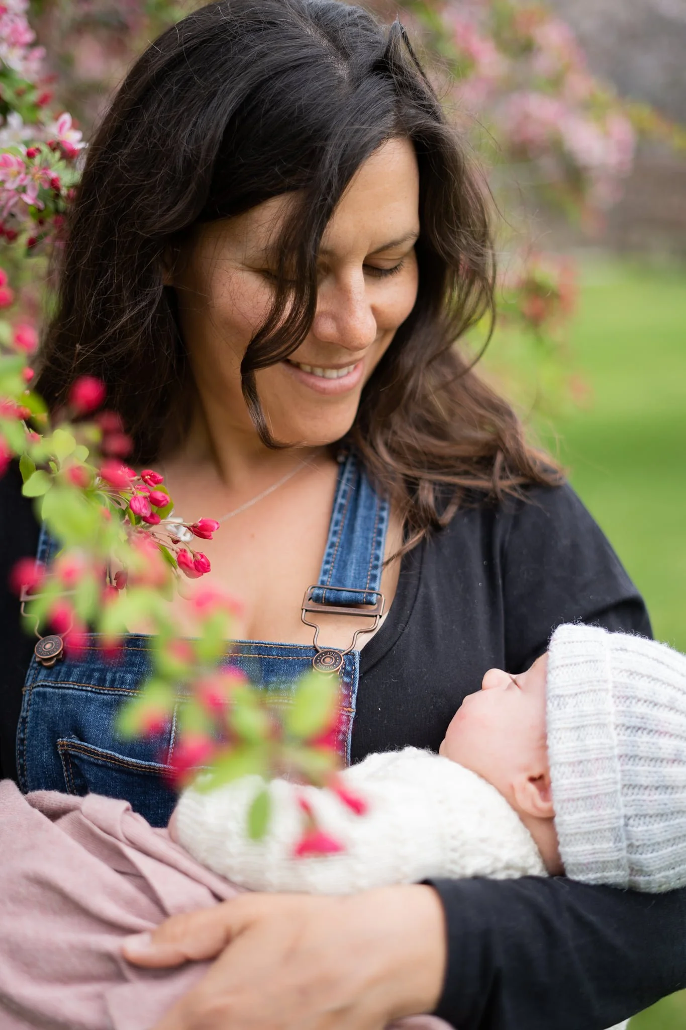 A woman holding a baby outdoors near pink flowers, both smiling and looking at each other.