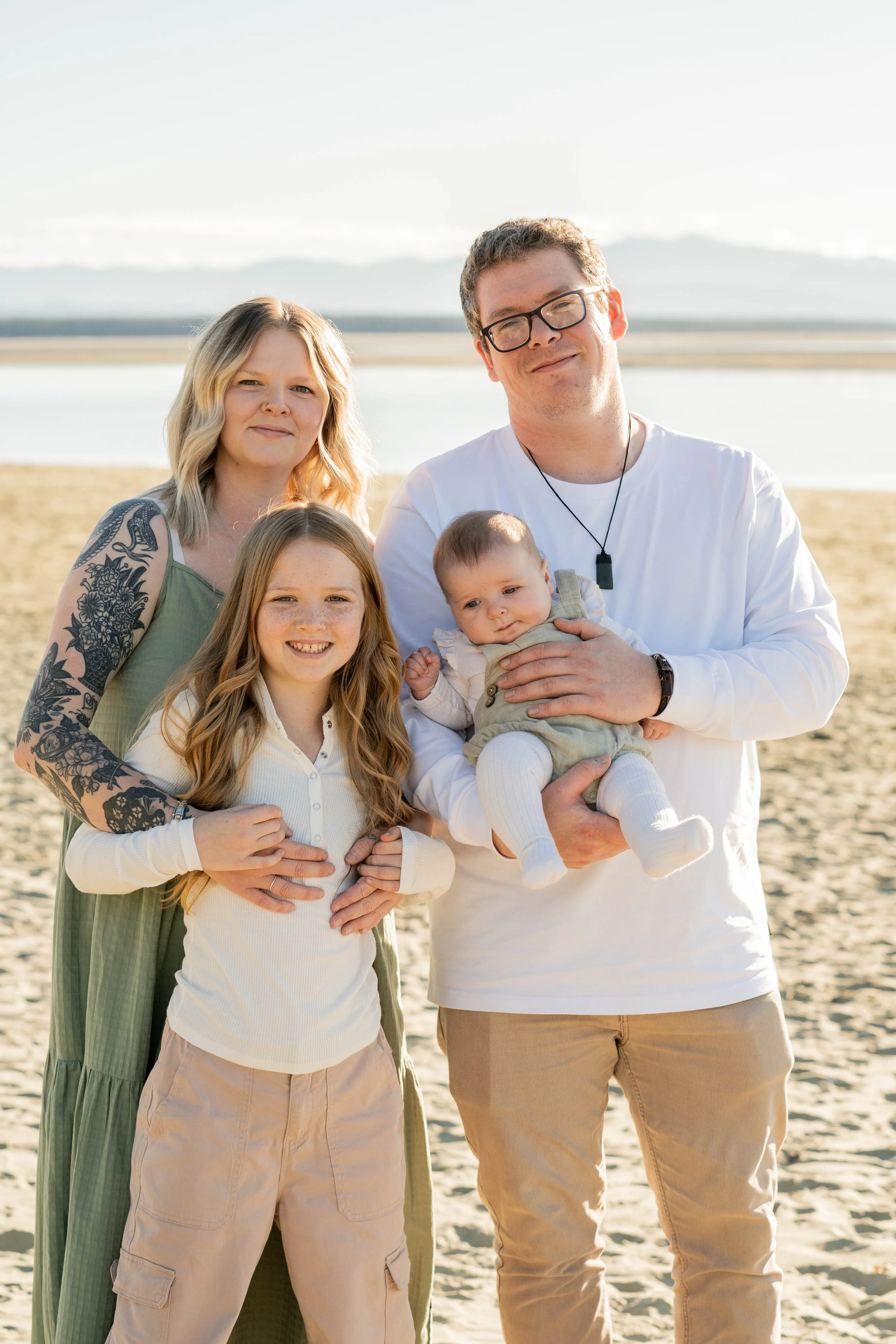 A family of four stands on a sandy beach, with a backdrop of water and mountains, on a sunny day. The father holds a baby, while the mother and a young girl stand beside them, smiling at the camera.
