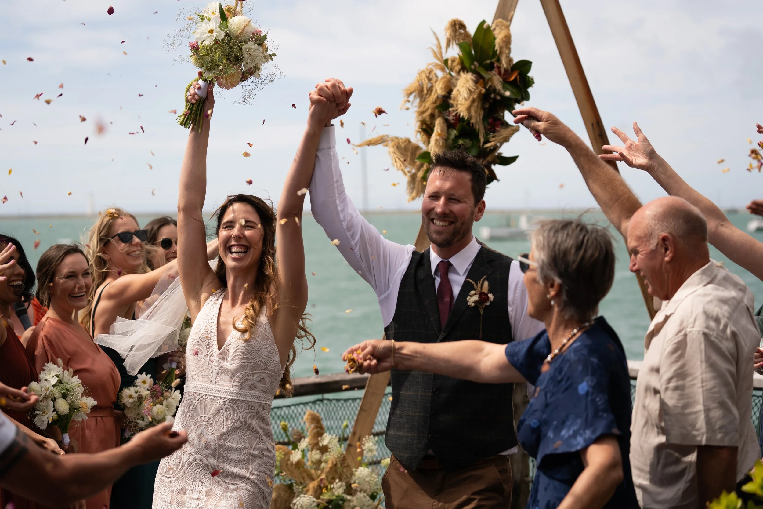 A joyful bride and groom celebrating with friends and family at a wedding outdoors by the water, throwing flower petals.