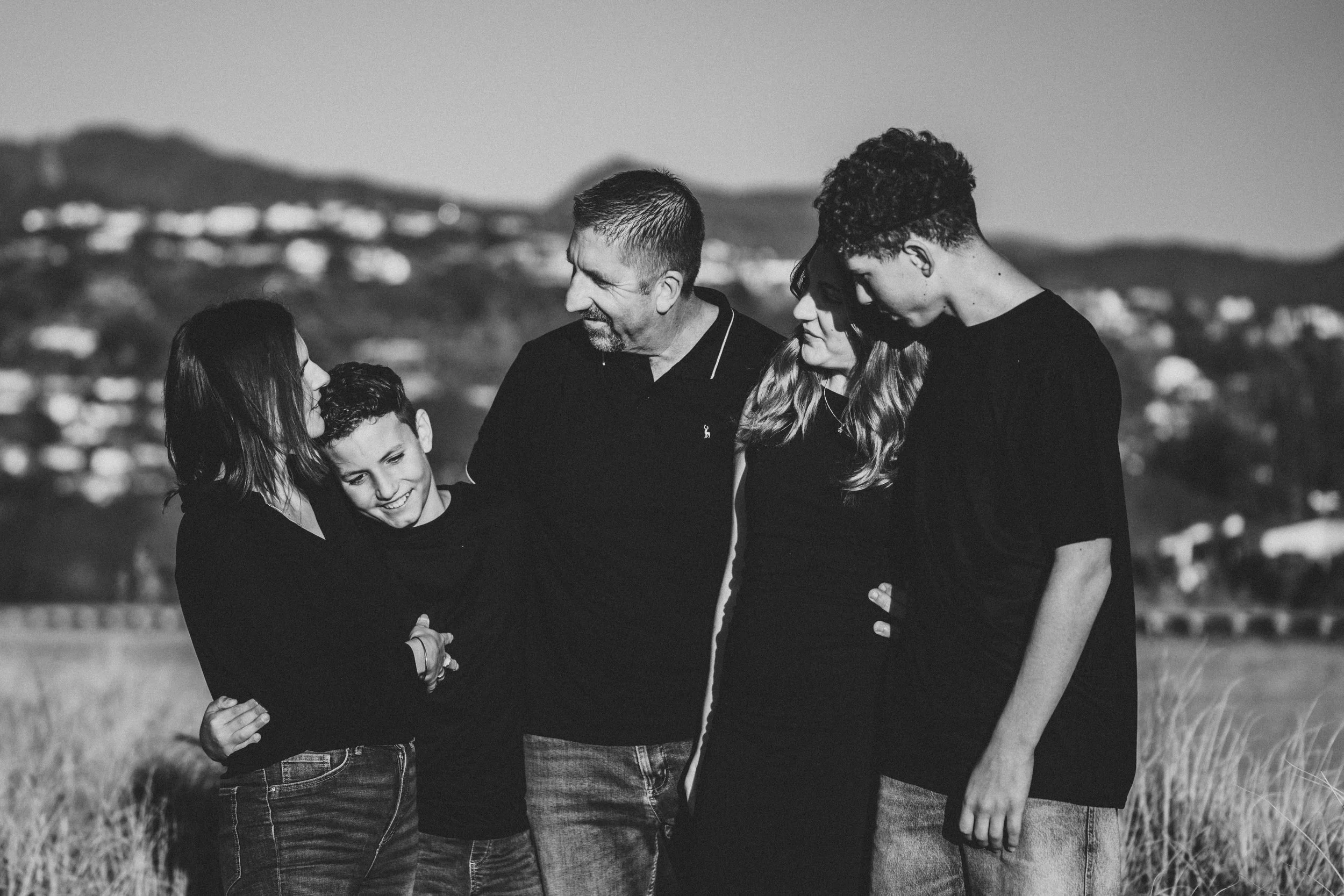 A family of six in a field outdoors, hugging and smiling at each other, with hills and houses in the background, in black and white.