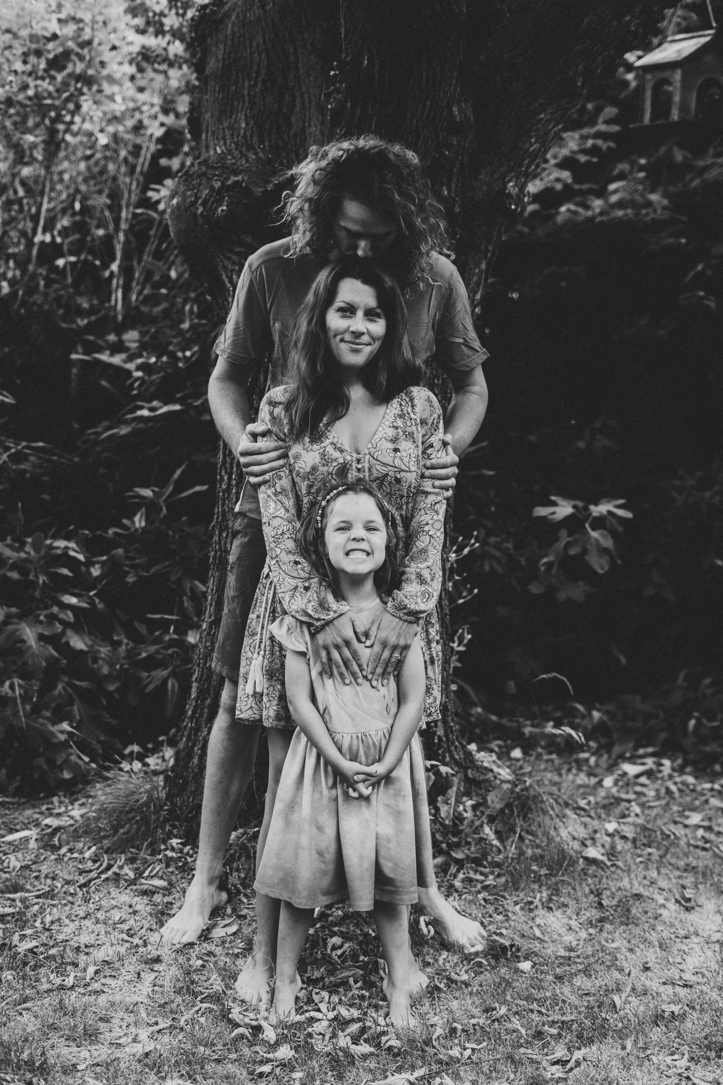 Black and white photo of three generations of females standing outdoors in front of a large tree. An adult woman is in the middle with an adult man behind her, and a young girl in front, all smiling and holding hands.