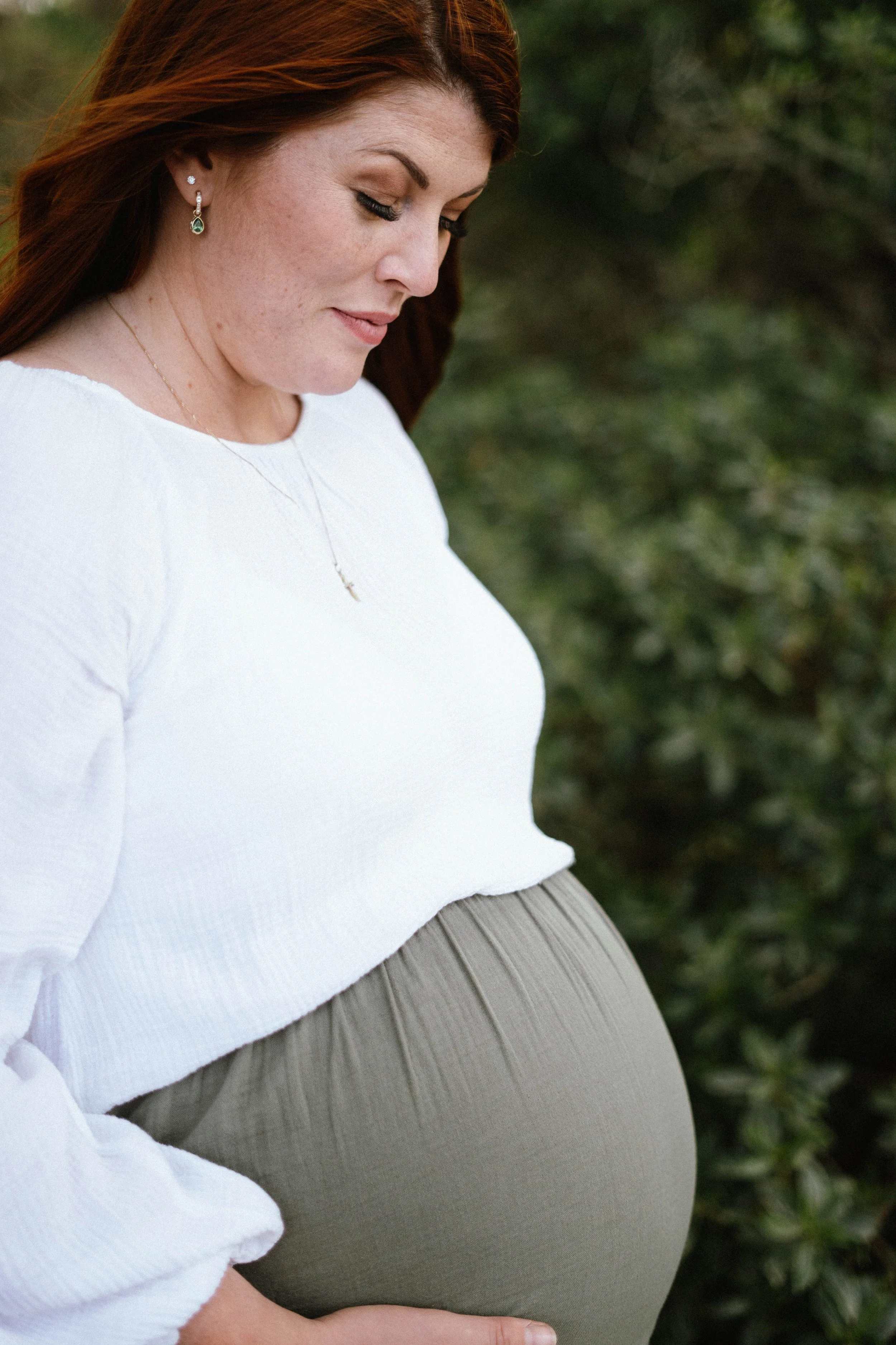 Pregnant woman with red hair and earrings in an outdoor setting, gently touching her large belly.