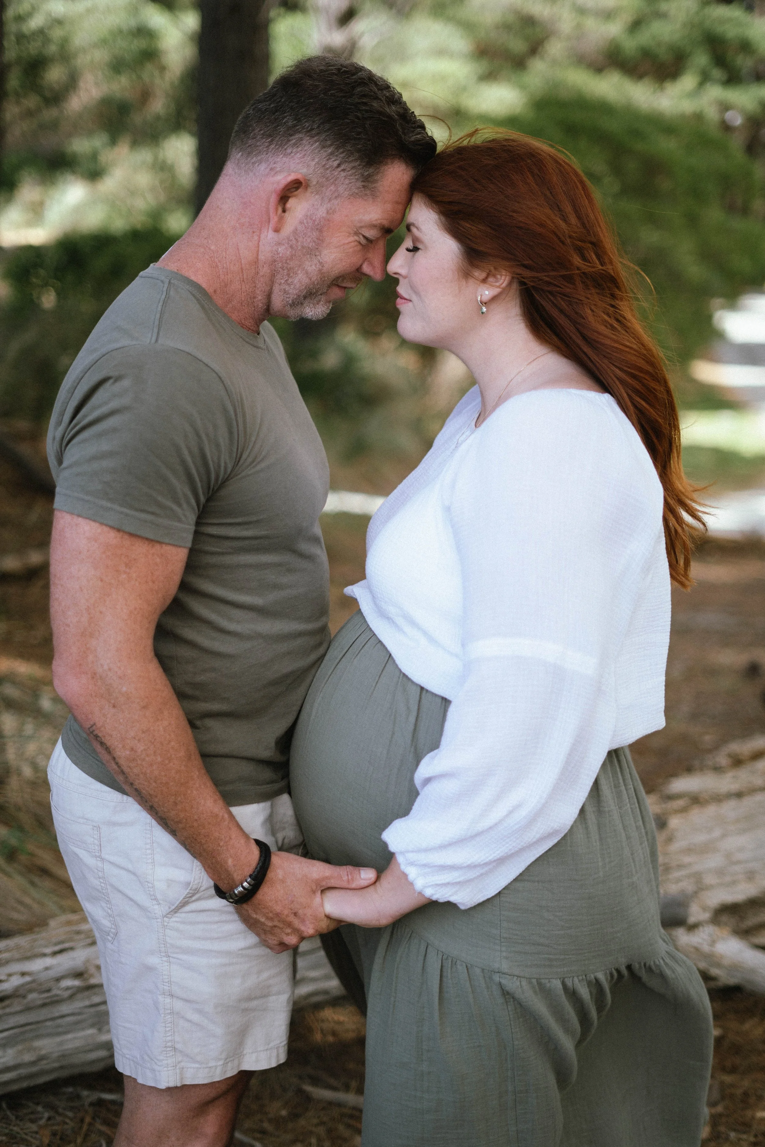 A pregnant woman with red hair and a man with short brown hair and a beard, both with their foreheads touching, holding hands in an outdoor setting with trees.