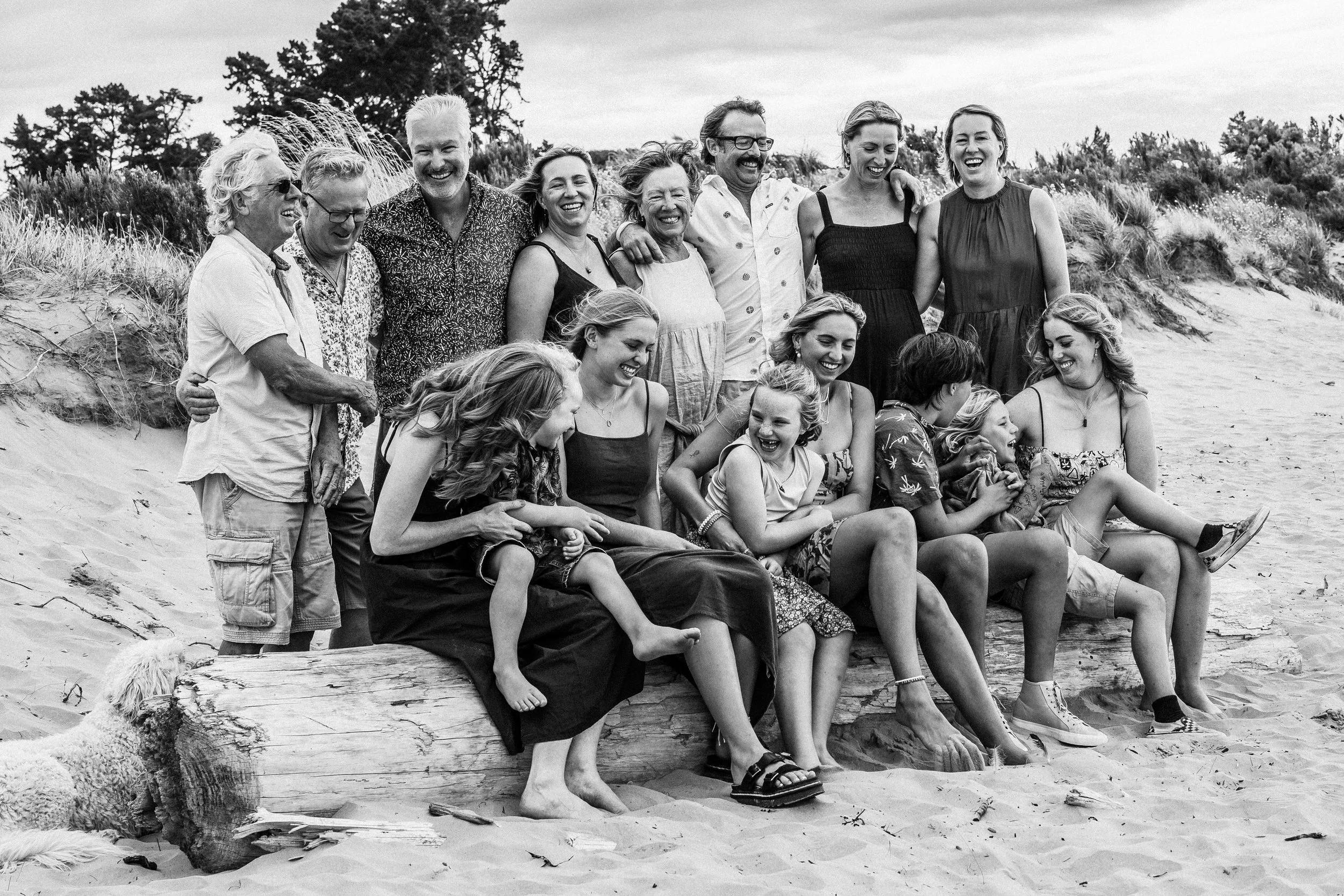 A group of people, including children and adults, sitting and standing on a large log on the beach, smiling and laughing together.